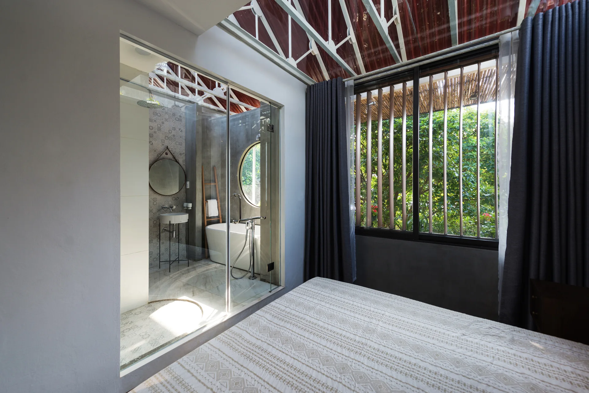 A contemporary bedroom features a bed in the foreground and a floor-to-ceiling glass partition separating it from an en-suite bathroom.