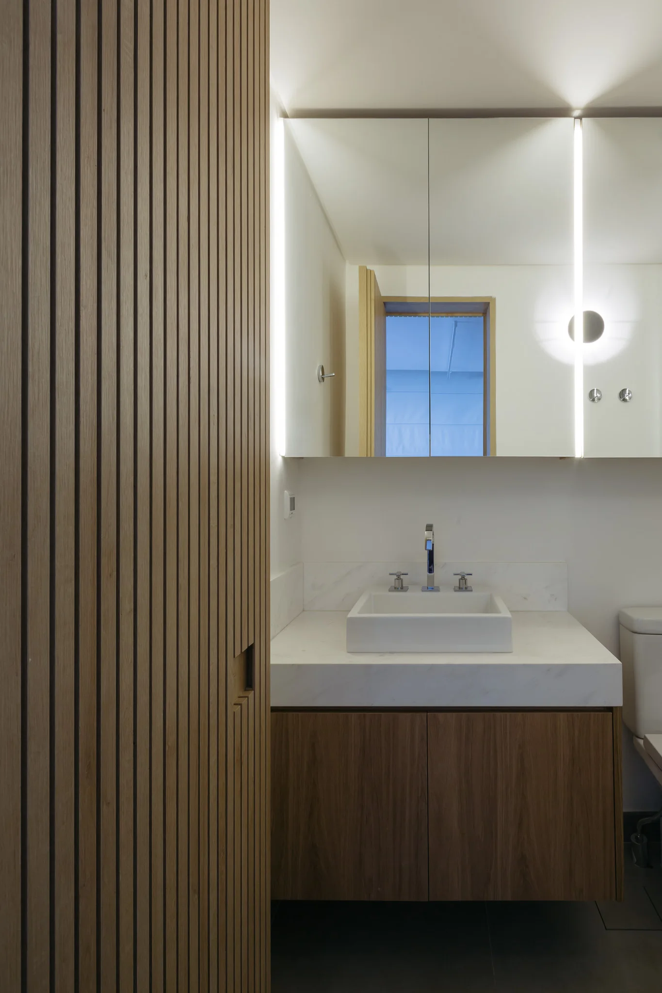 A modern bathroom vanity with a white marble countertop, a wood cabinet, and vertical slatted wood paneling next to a backlit mirror.