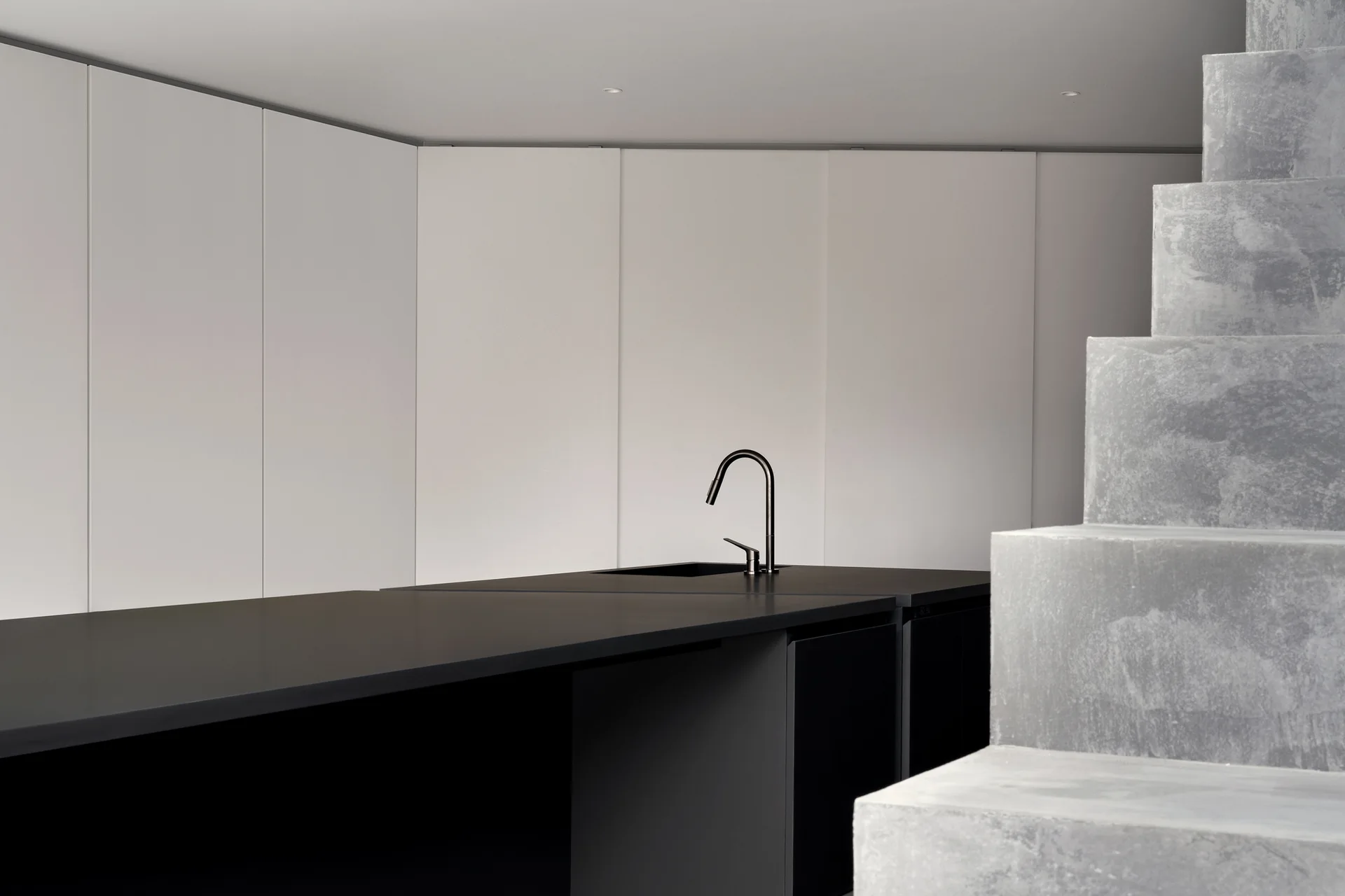 A minimalist kitchen features a matte black island with a black faucet set against a seamless white panel wall and concrete stairs.