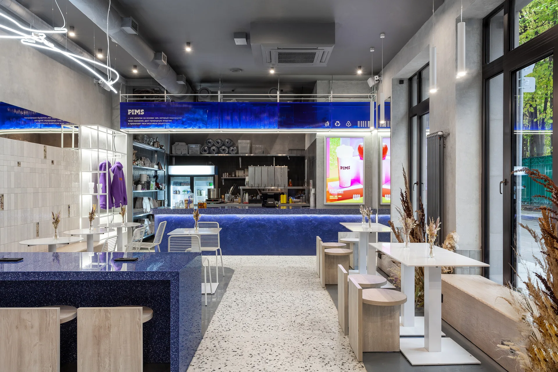 A modern cafe interior features a bright blue speckled bar counter, terrazzo flooring, light wood stools, white square tables, and large windows.