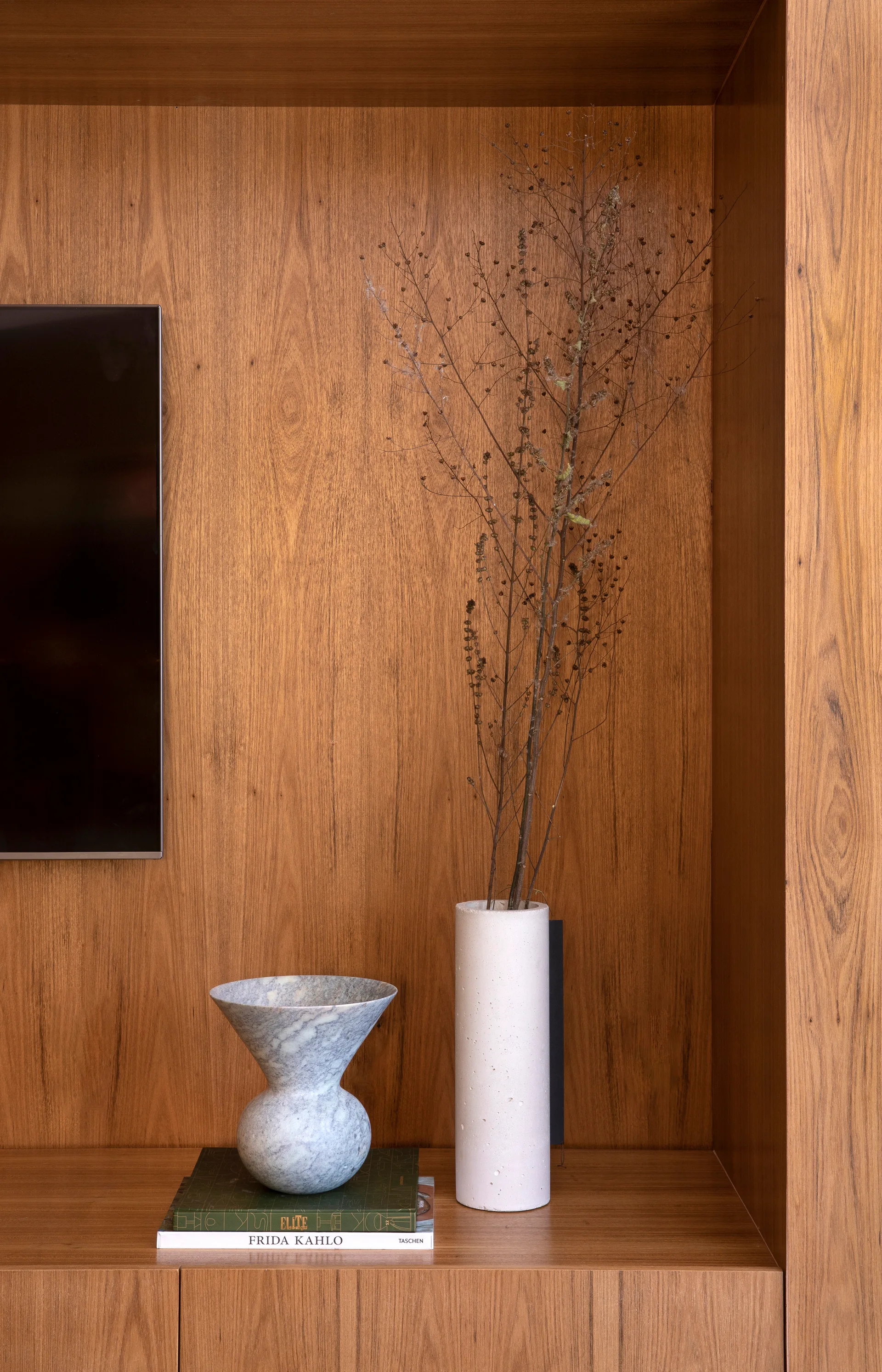 A close-up of a wood-paneled display niche featuring a marble bowl on stacked books, a tall white vase with dried branches, and a TV edge.