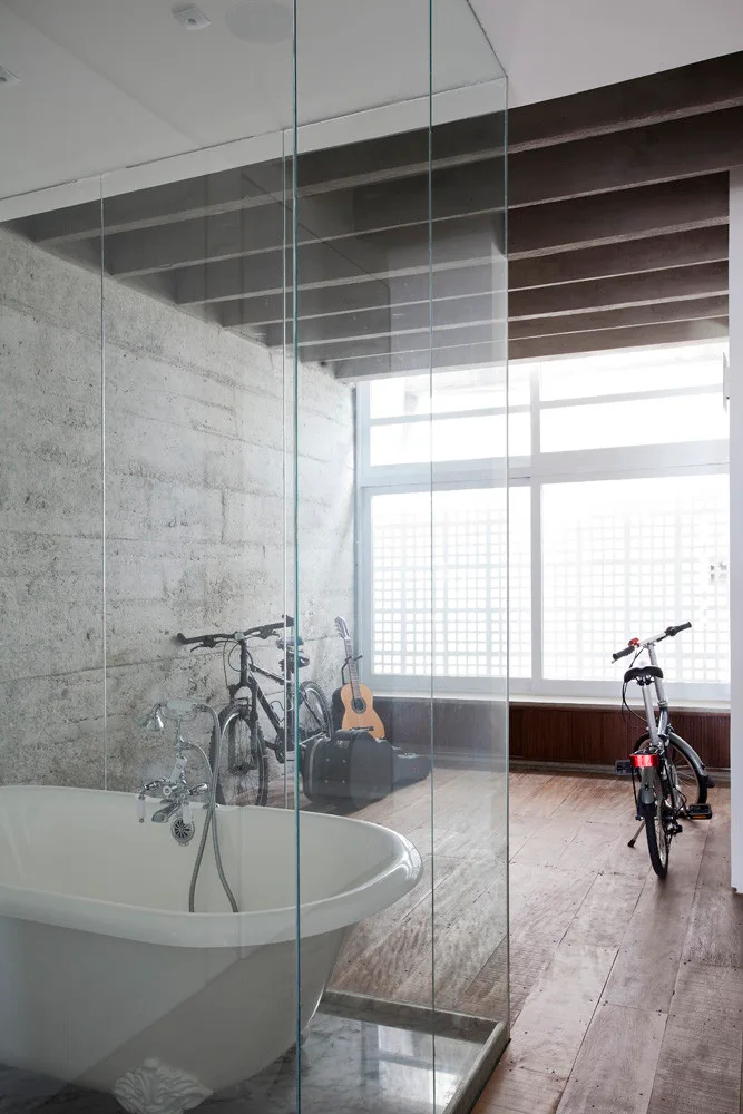 An industrial-style bathroom features a white clawfoot bathtub enclosed by a glass partition next to a textured concrete wall and wood floor.