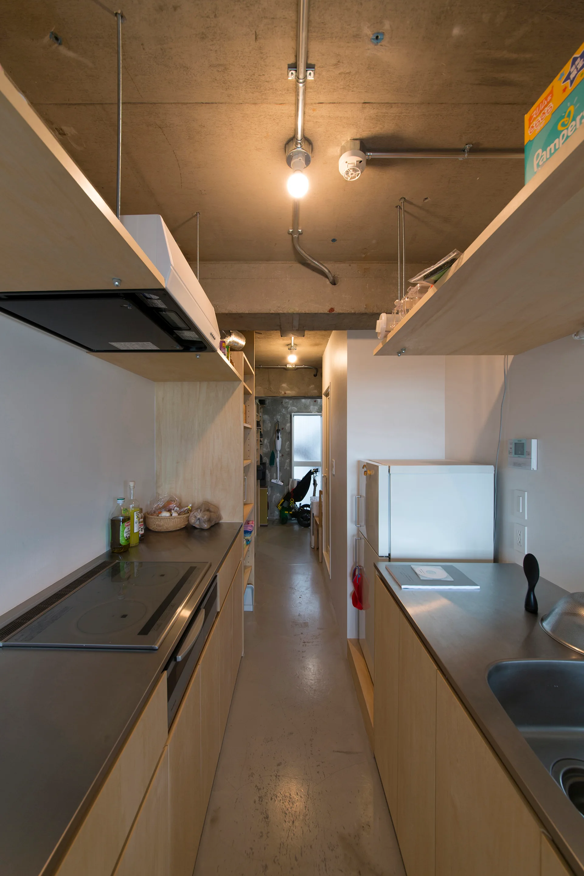 A narrow galley kitchen features light wood cabinets, stainless steel countertops, and an exposed concrete ceiling with metal conduits.