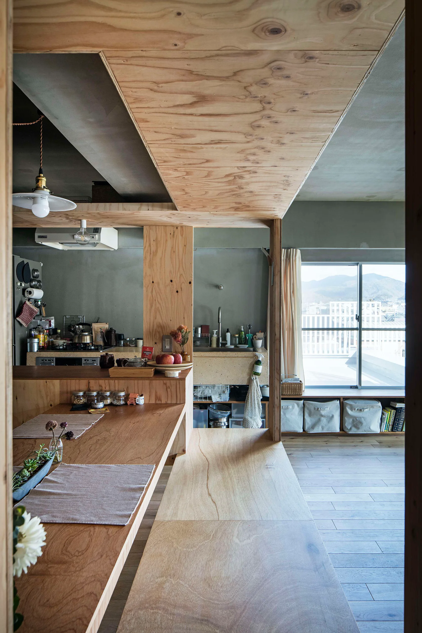 A minimalist kitchen features natural plywood cladding on the ceiling and structural partitions, with a wooden island and sage green walls.