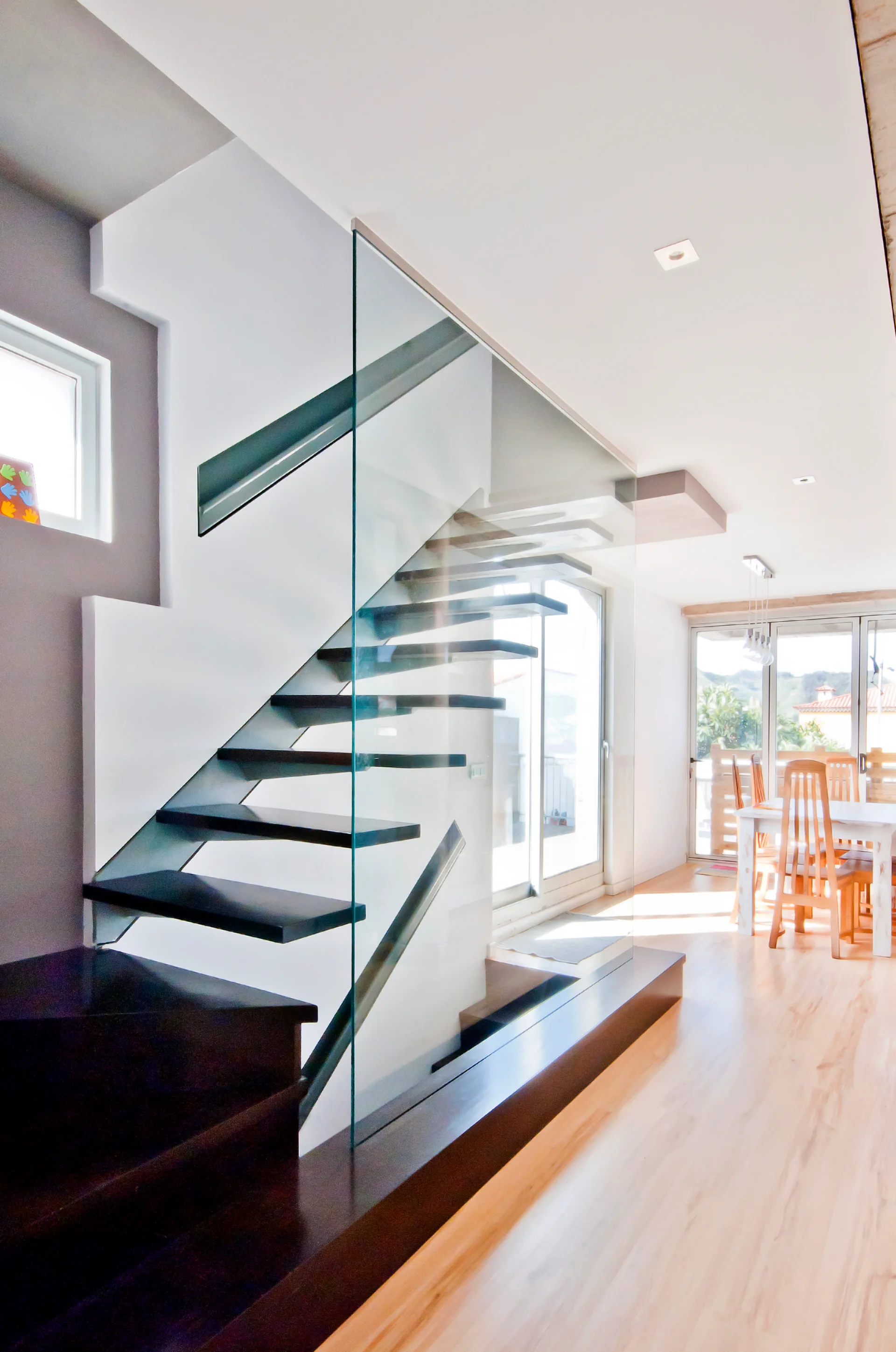 A modern staircase with dark wood treads, open risers, and a clear glass railing stands in a bright residential interior with light wood flooring.