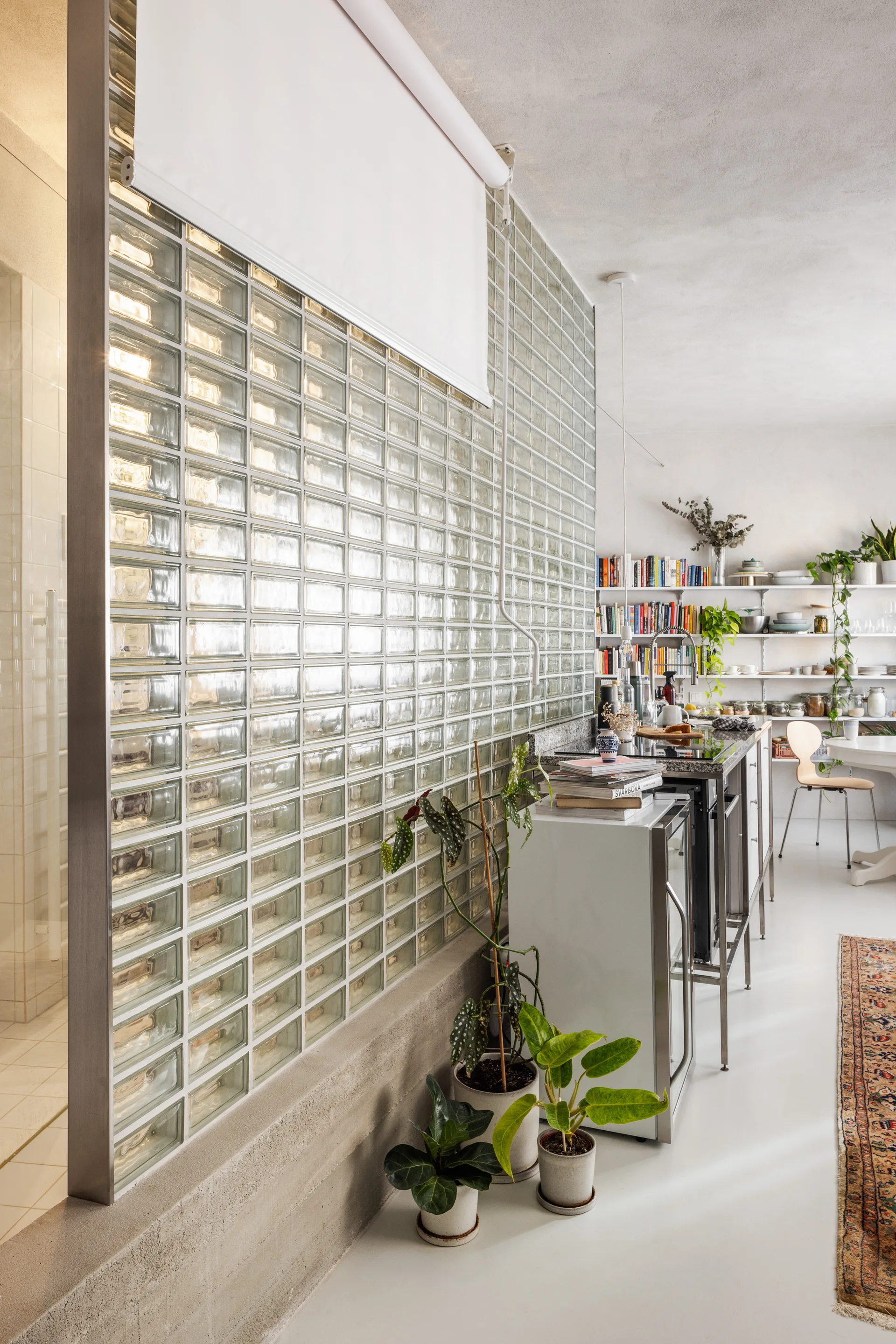 A bright kitchen interior featuring a large glass block partition wall on a concrete base next to a metal kitchen island and open shelving.