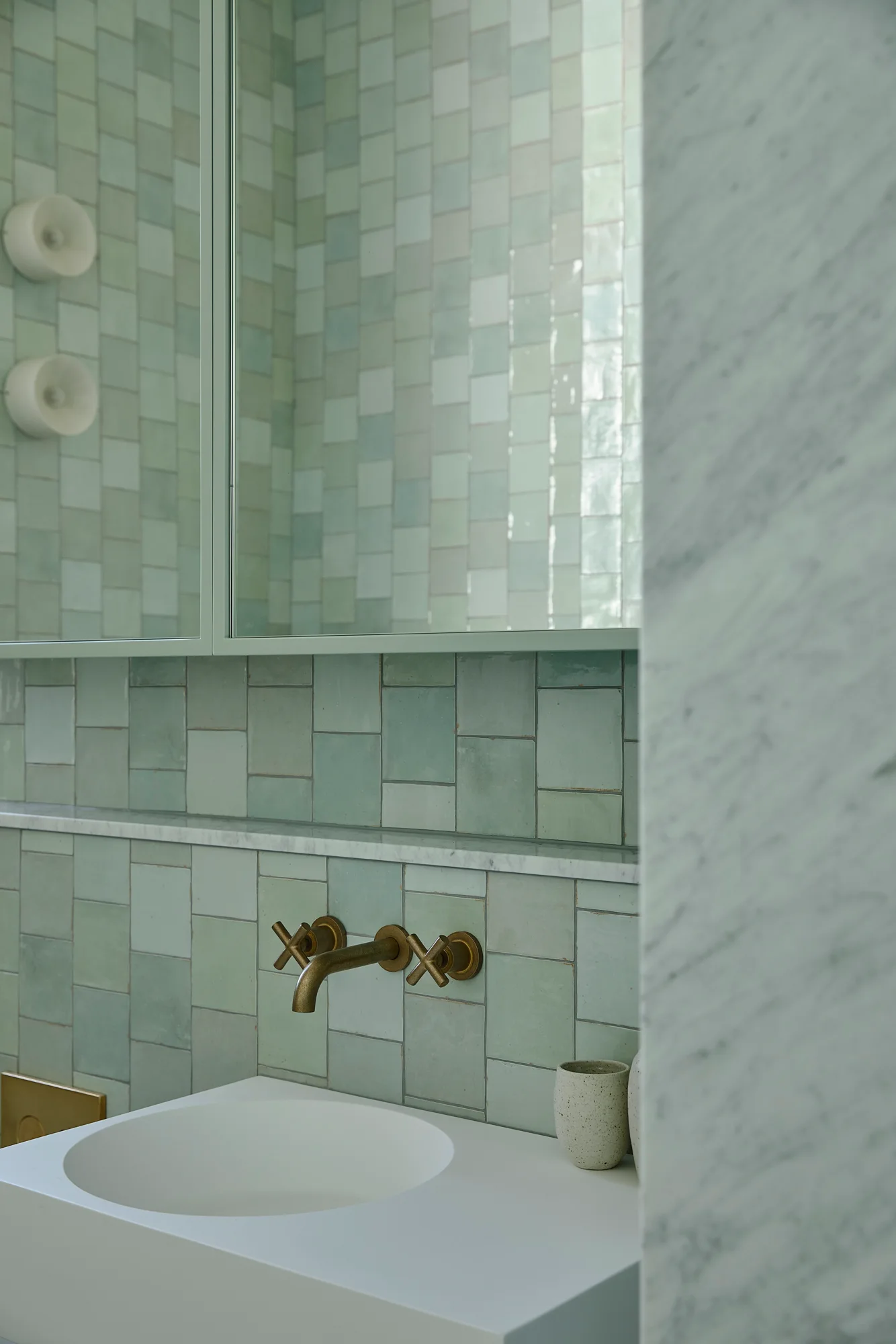 A modern bathroom vanity features a white integrated sink, a wall-mounted brass faucet, sage green tiled walls, and a marble partition.