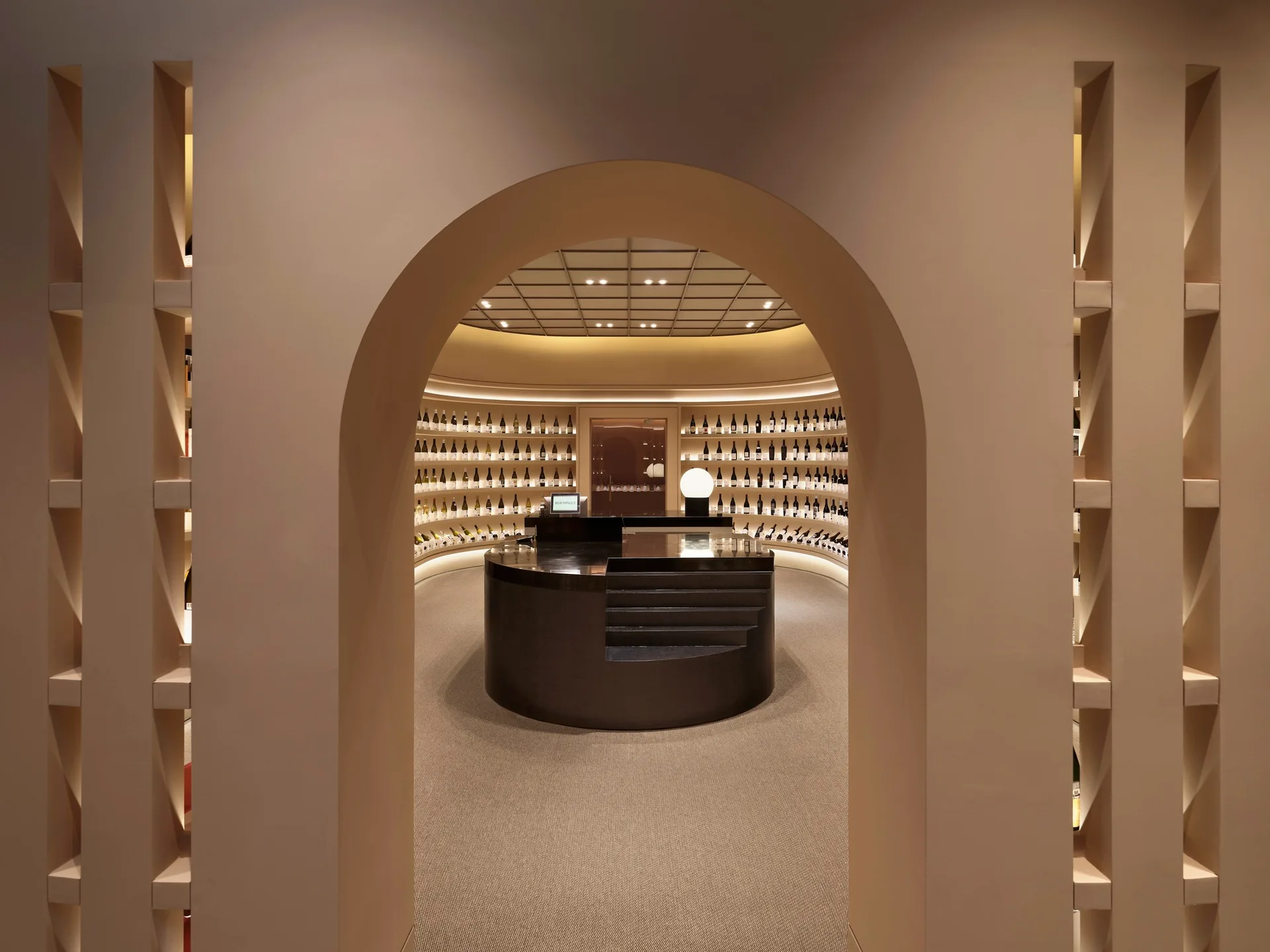 A circular wine display room with curved wall shelving seen through a large beige arched entryway.