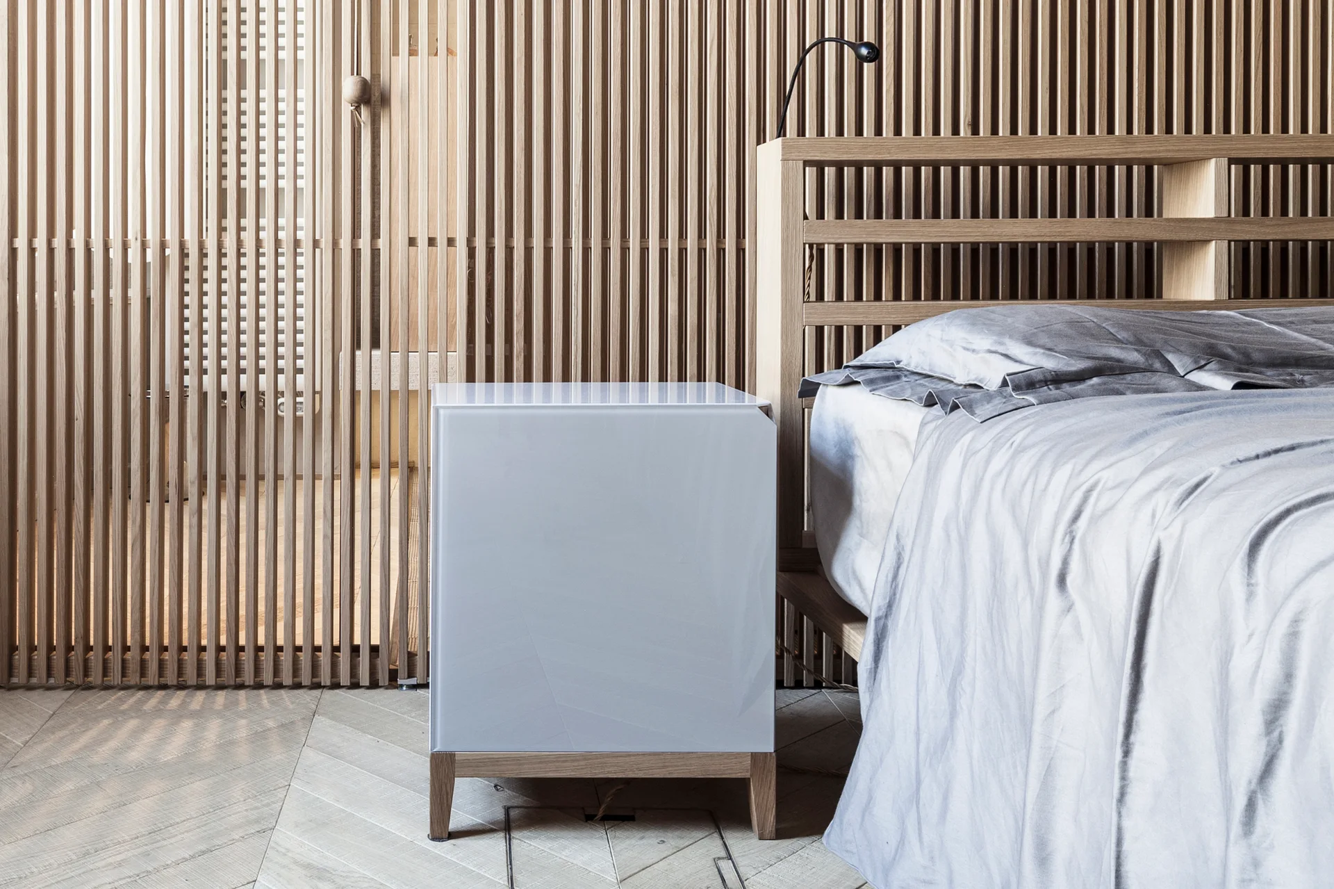 A minimalist bedroom features a bed with grey linens, a glossy white cube nightstand, and a light wood slatted wall behind the headboard.