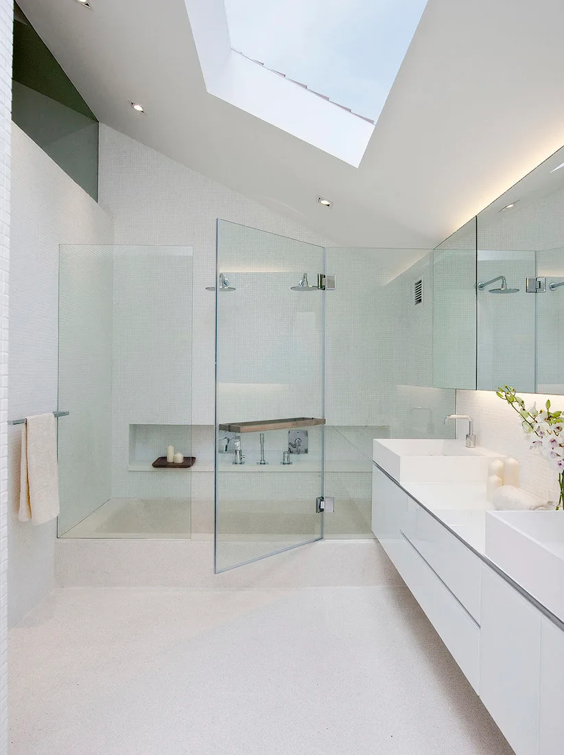 A modern white bathroom features a floating double vanity, a glass-enclosed walk-in shower, and a large skylight in the sloped ceiling.