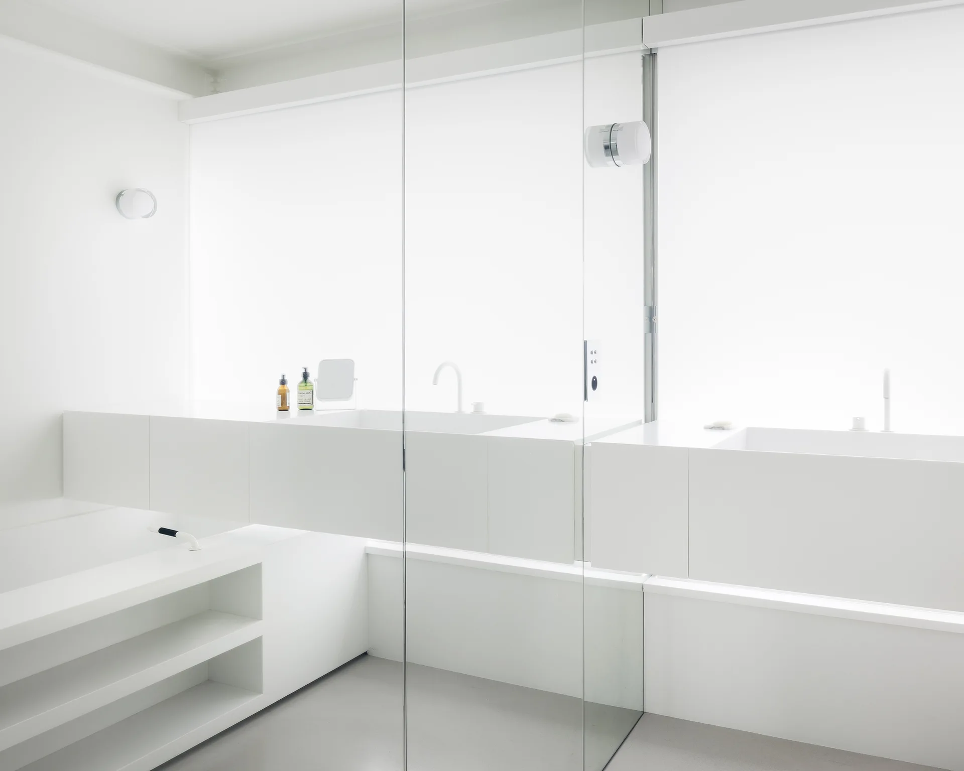 A minimalist white bathroom features a floating vanity, matte white fixtures, a glass partition, and a light grey floor under diffused window light.