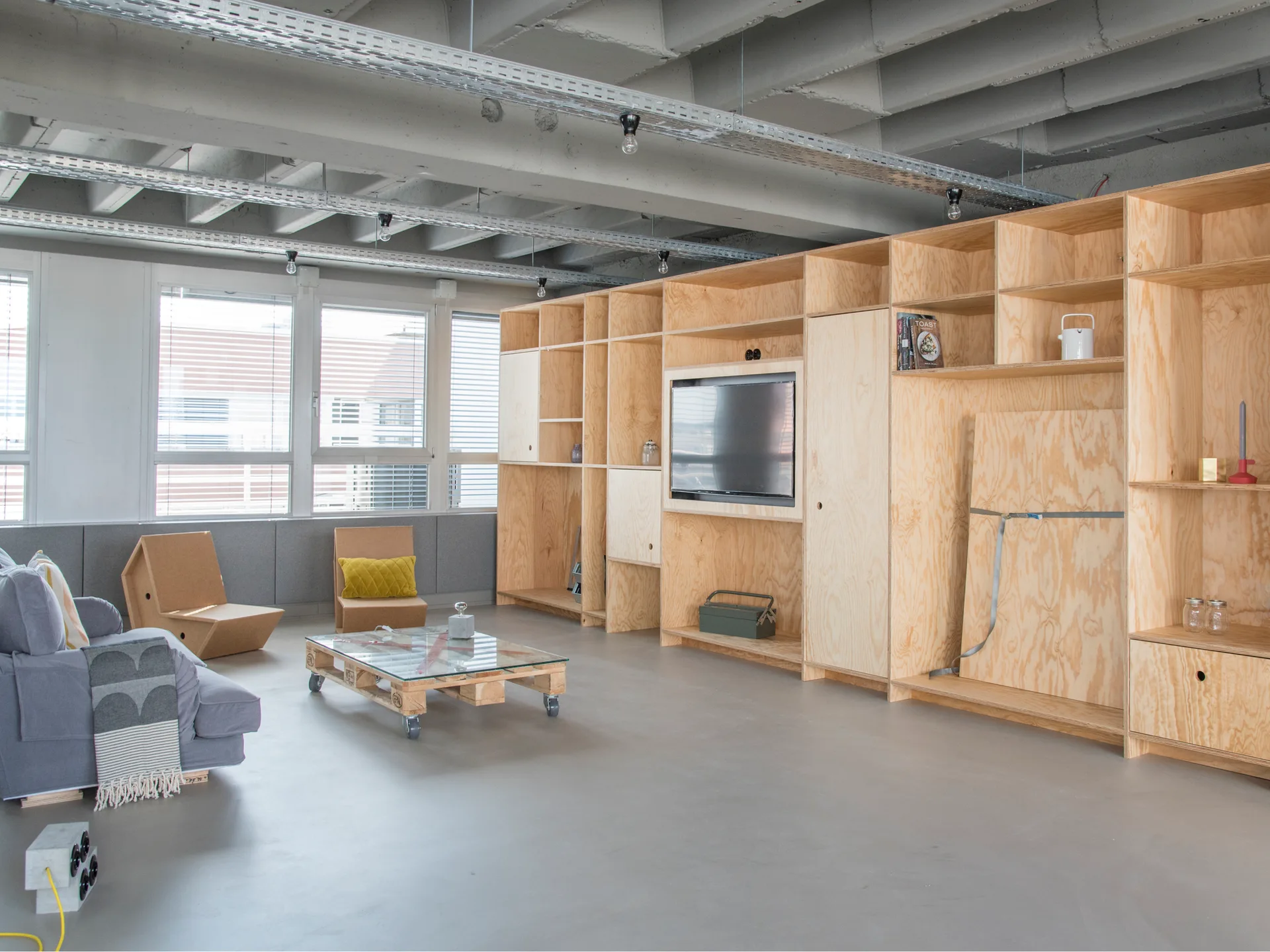 An industrial living room features a large custom plywood wall storage unit, a grey concrete floor, exposed ceiling beams, and a low pallet coffee table.