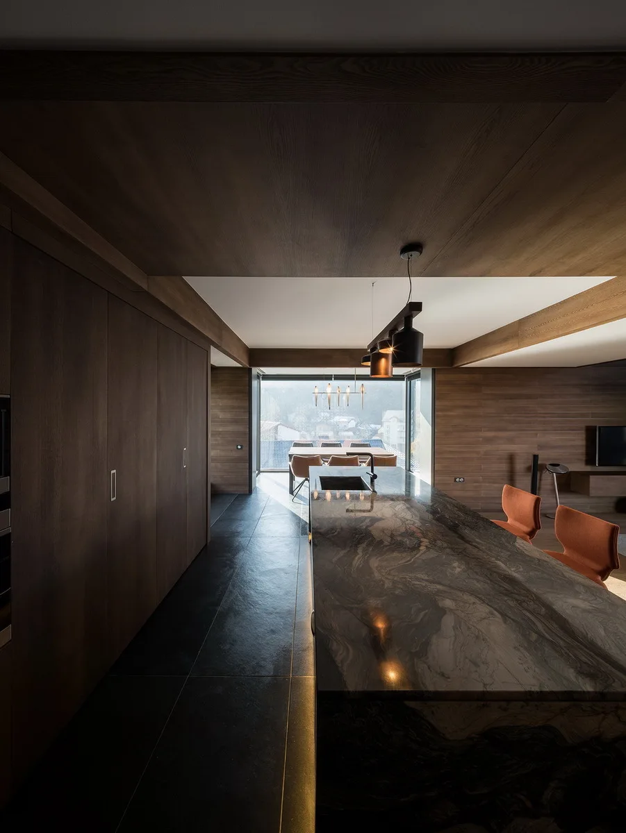 A modern kitchen featuring a dark veined stone island, matte wood-paneled walls and ceiling, dark tile floors, and orange accent chairs in the background.