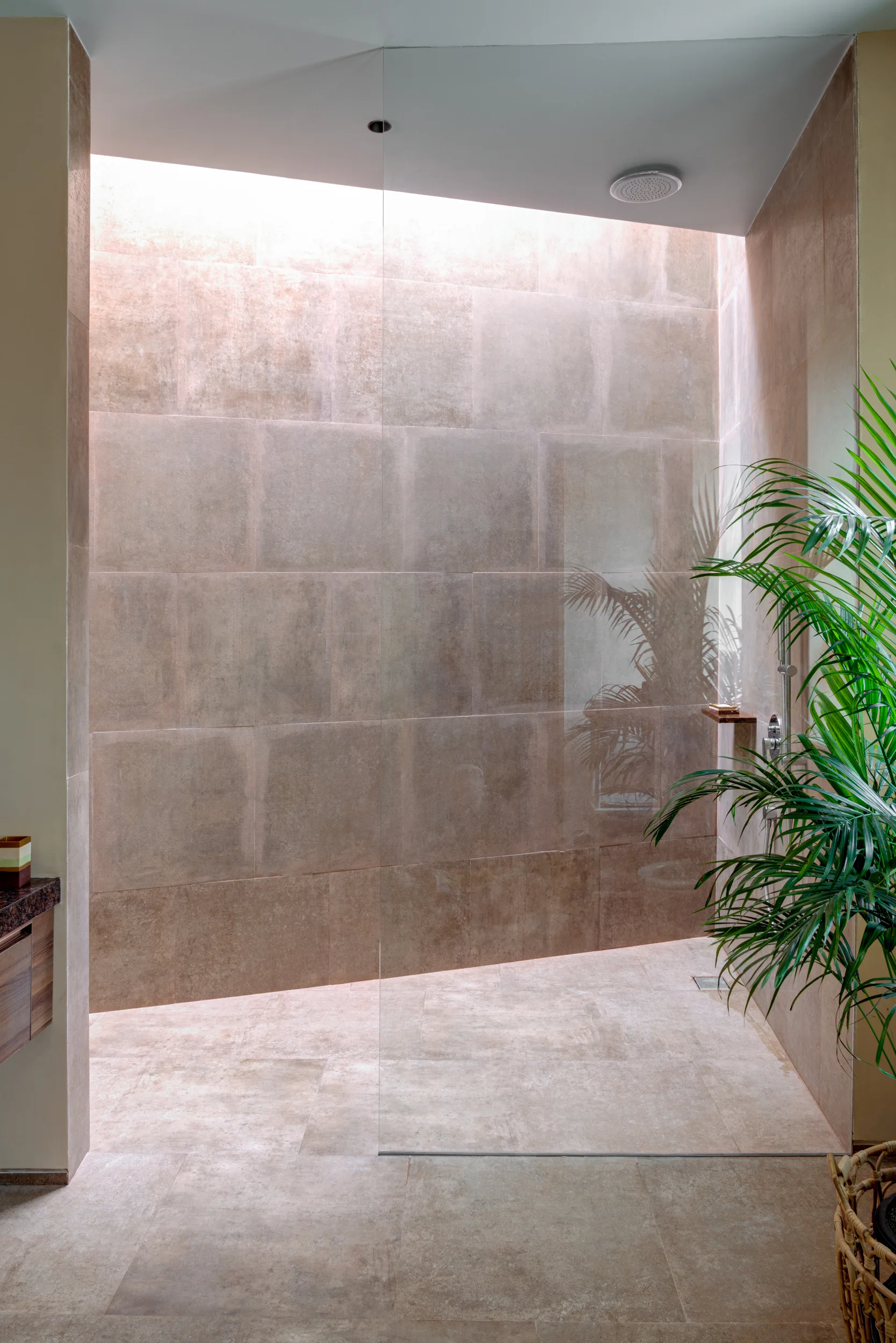 A modern walk-in shower with large-format beige tiles, a floor-to-ceiling glass partition, a ceiling shower head, and a natural light well above.