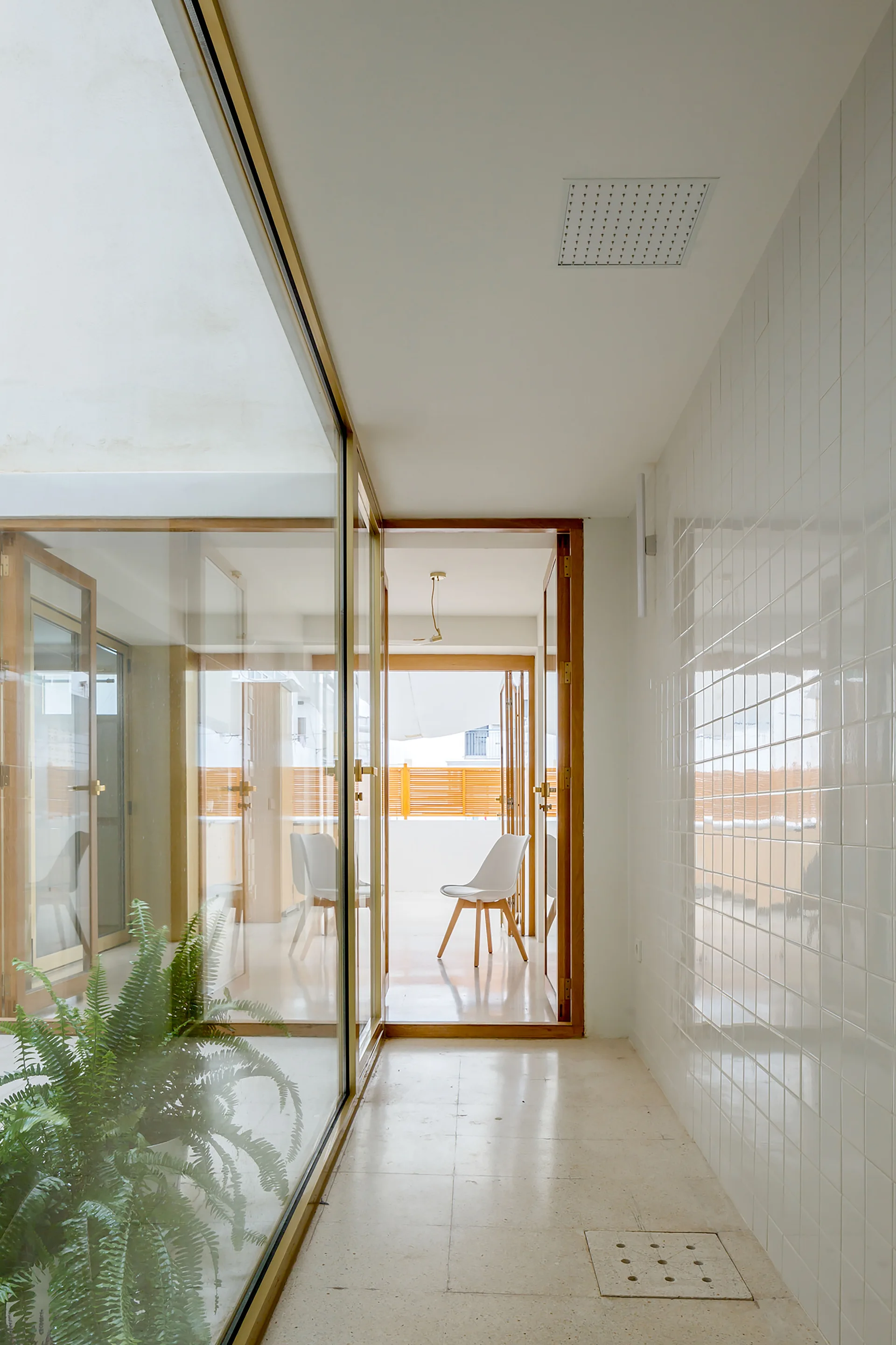 A bright, minimalist corridor featuring white glossy tiled walls, a ceiling shower head with a floor drain, and glass walls looking out to a terrace.