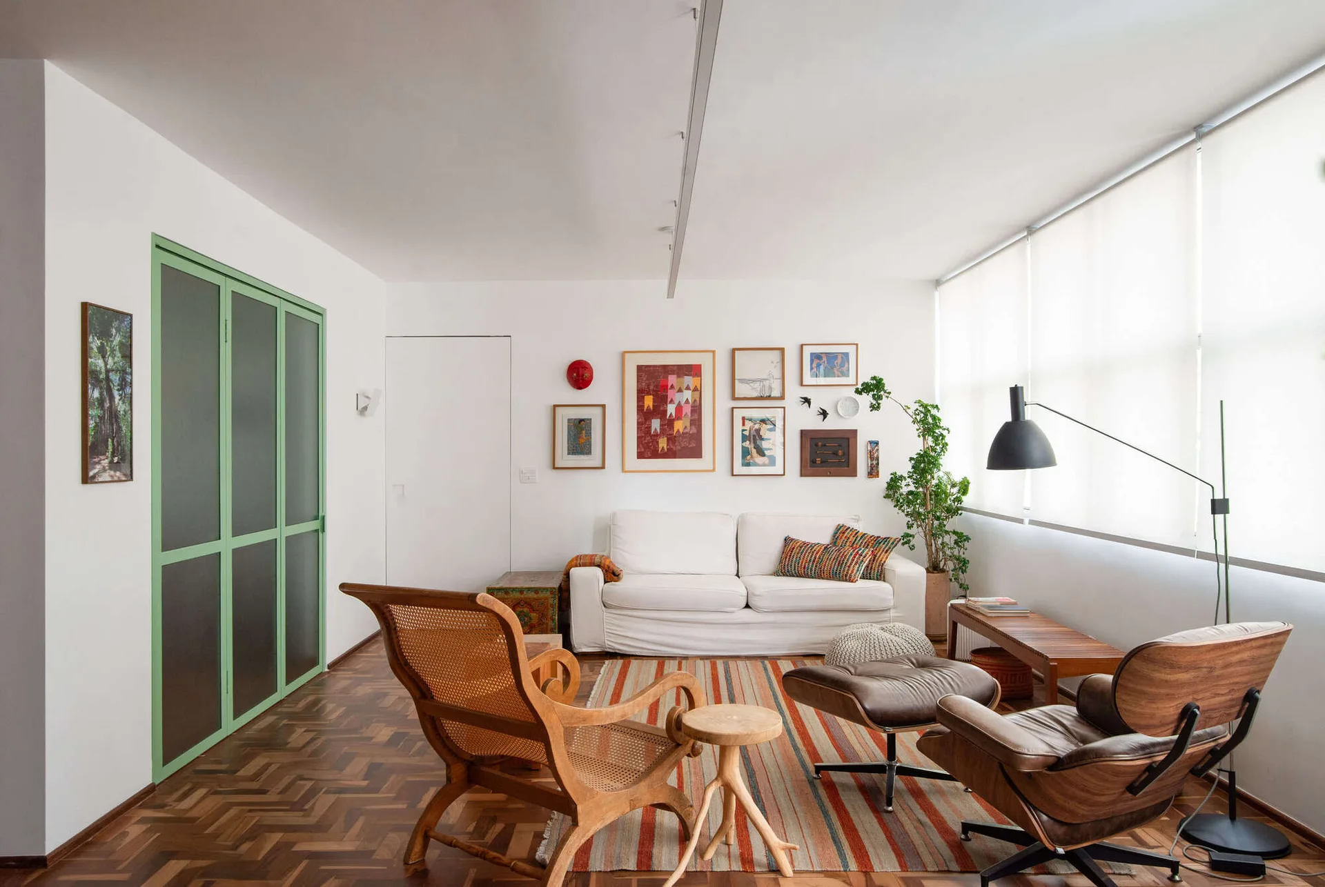 An eclectic living room with a white sofa, a green-framed sliding door, a striped area rug, and mid-century wooden lounge chairs on herringbone floors.
