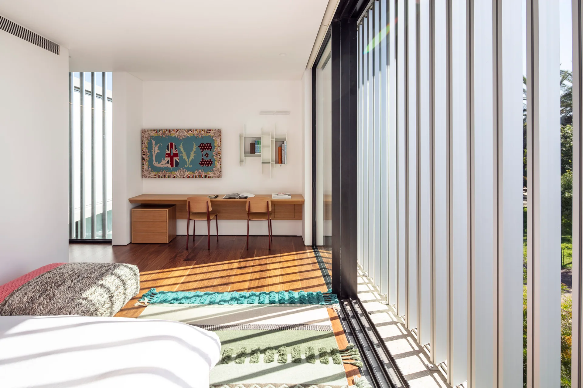 A modernist bedroom featuring a built-in wood desk with two chairs, a textured rug on wood floors, and vertical glass louvers casting striped shadows.