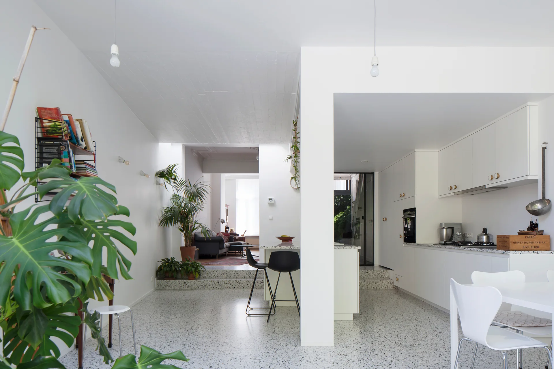 A modern open-plan kitchen featuring white cabinets, a dining table, black bar stools, and a multi-level speckled terrazzo floor leading to a living room.