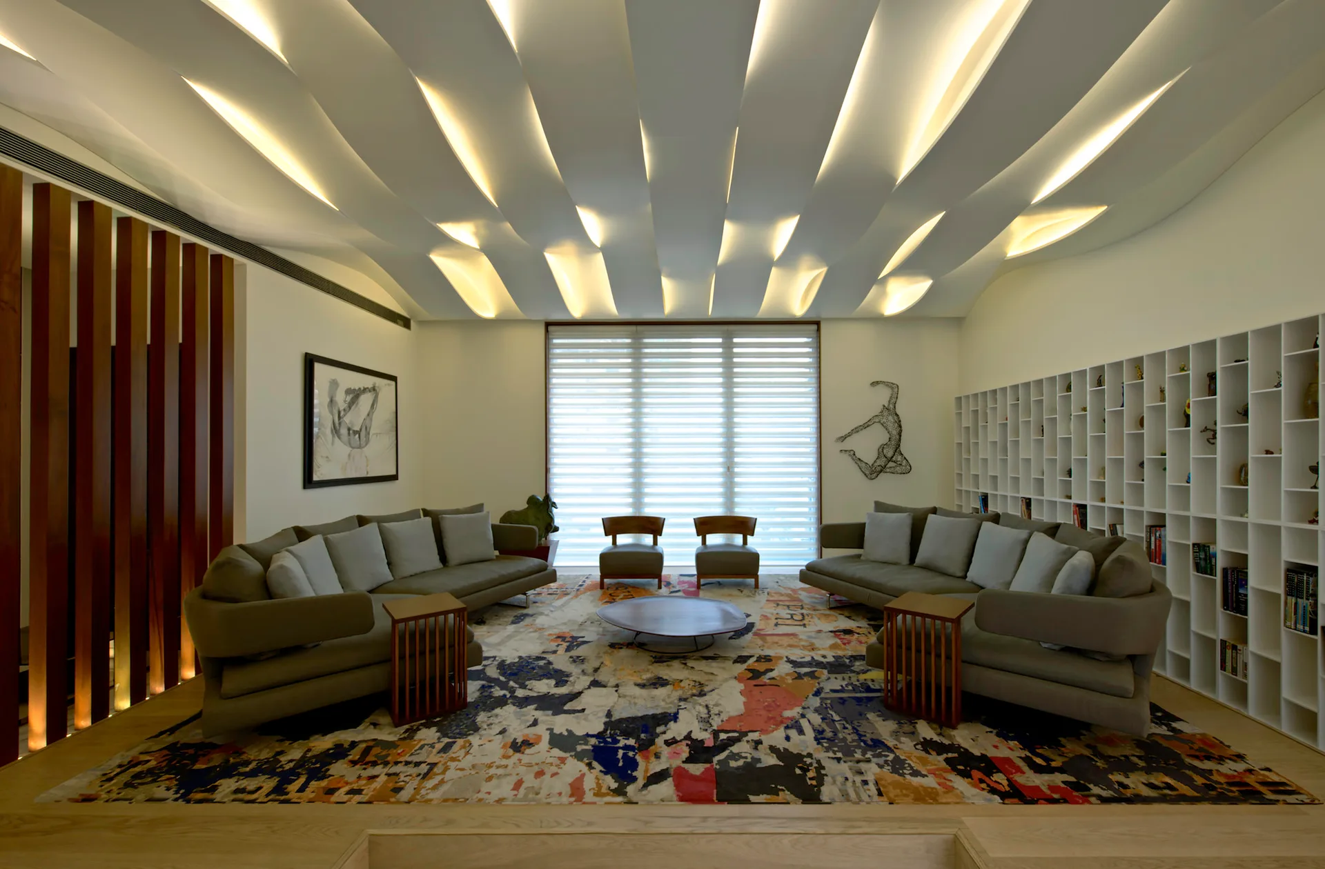 A symmetrical living room featuring two grey sofas on an abstract patterned rug beneath a sculptural wave-like ceiling with warm recessed lighting.