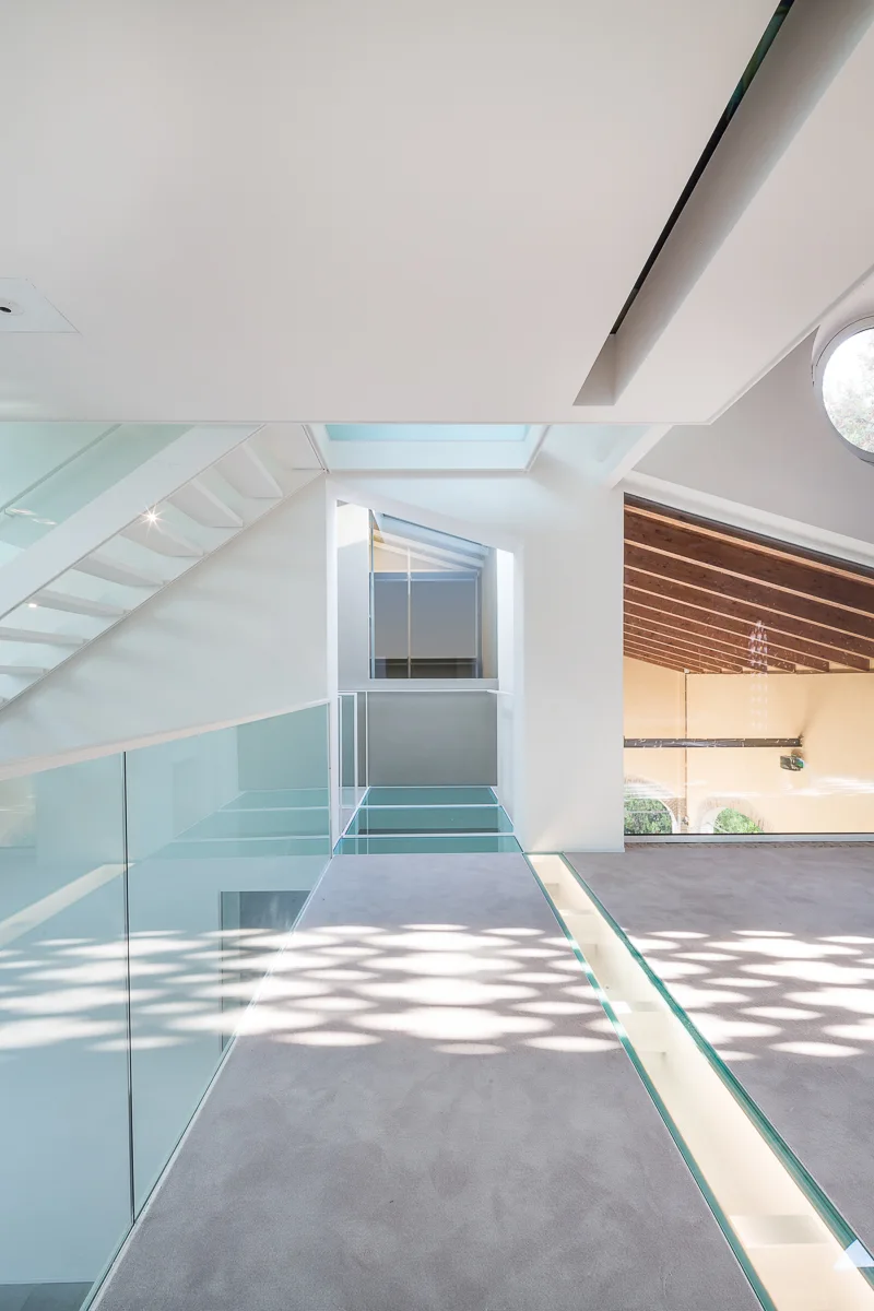 A modern white interior landing features a glass-railed staircase, a carpeted walkway with glass floor inserts, and natural light from a skylight.