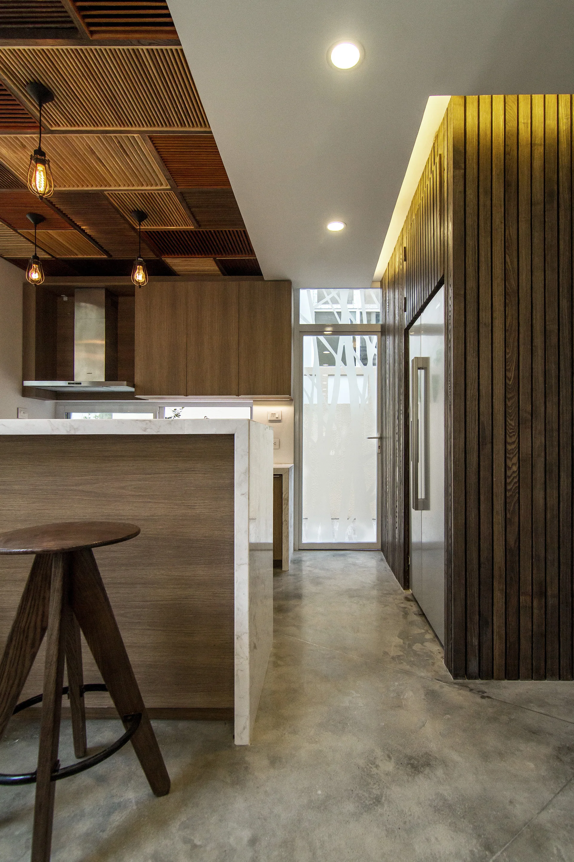 A modern residential kitchen featuring a slatted wood ceiling, a stone-topped island, vertical wood-paneled walls, and polished concrete flooring.