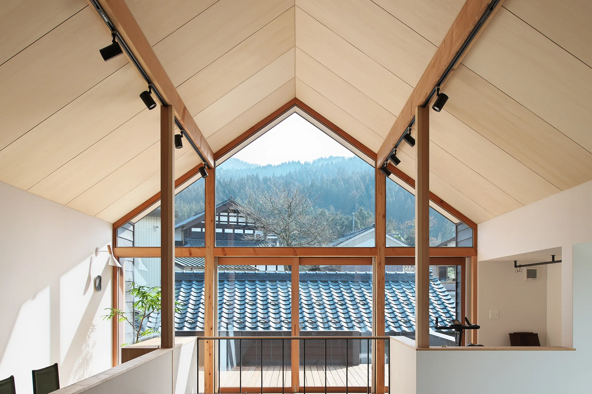 An airy loft interior featuring a vaulted wood plank ceiling with black track lights and a large peak window overlooking a forested hill.