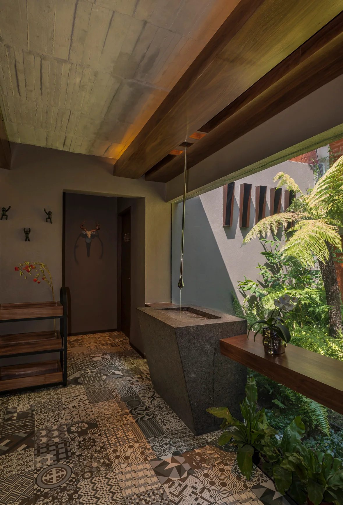 A stone water basin sits near a large window overlooking green plants, set against dark walls and a black-and-white patterned tile floor.
