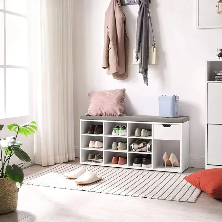 A white cubby shoe storage bench with a grey upholstered seat cushion is placed in a bright entryway next to a striped rug and wall hooks.