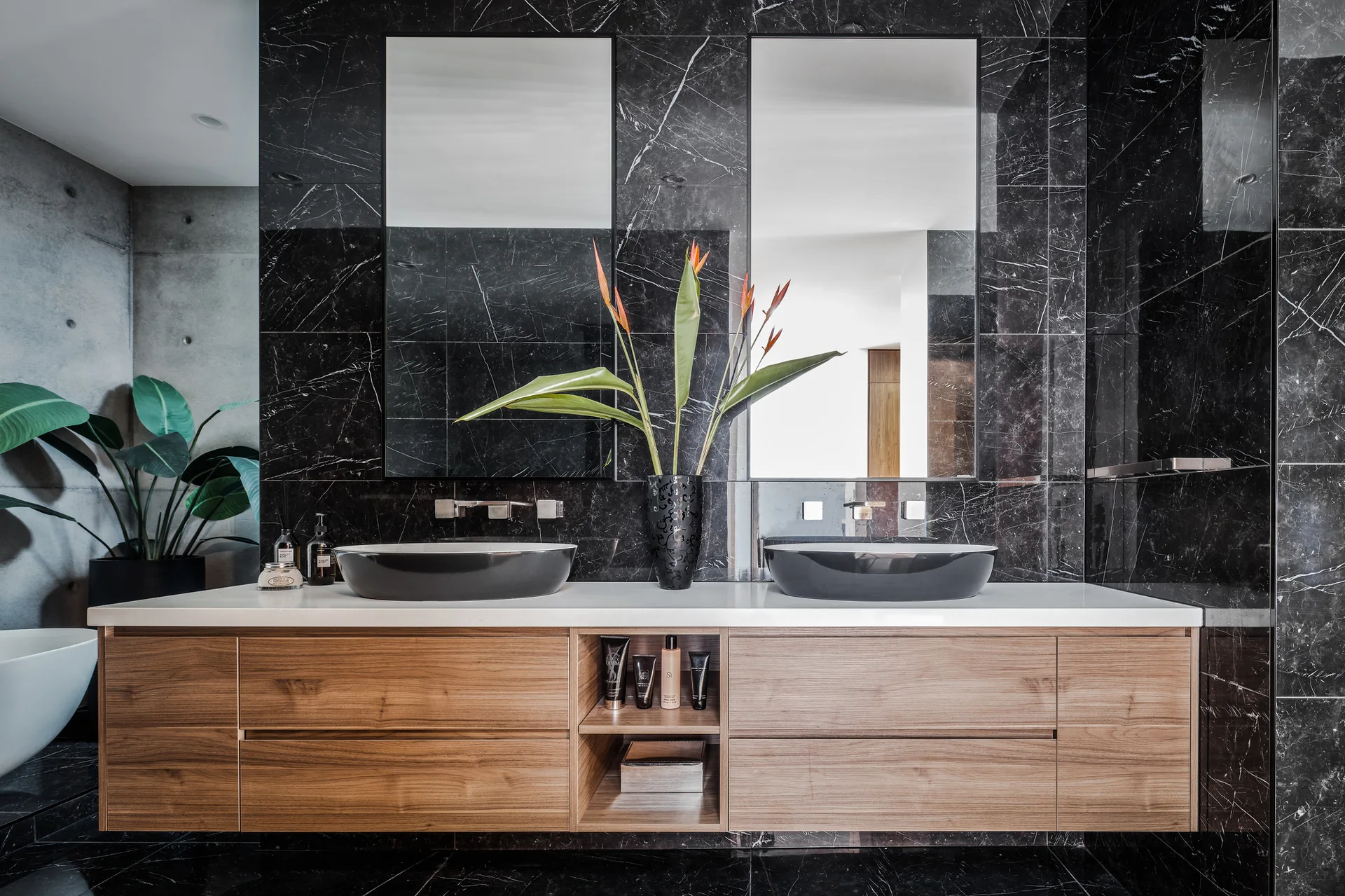 A modern bathroom featuring a floating wooden vanity with two black vessel sinks against a highly polished black marble wall with white veining.