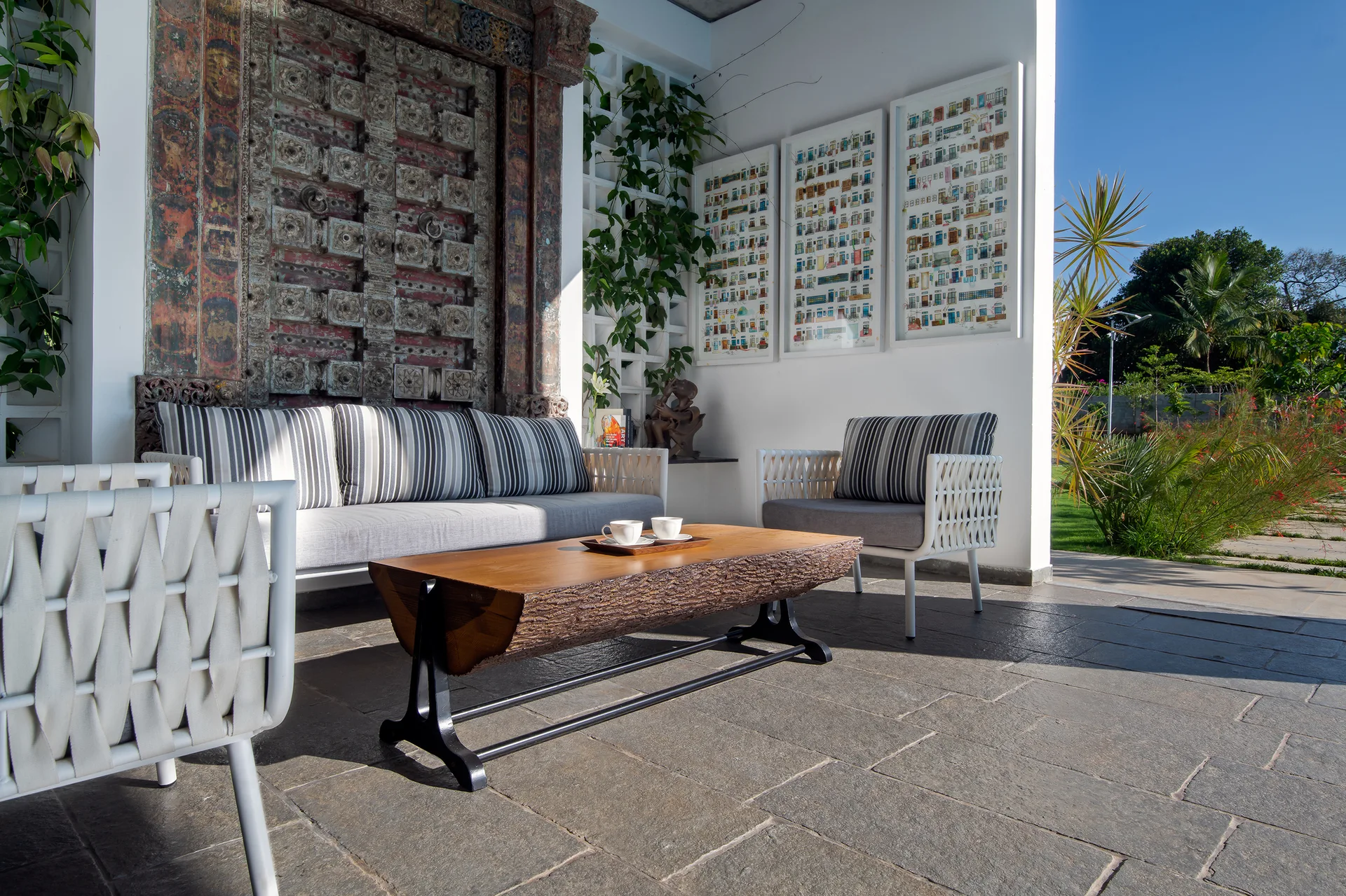 An outdoor patio featuring striped sofa seating, a live-edge wooden coffee table, and a large ornately carved wooden door on a white wall.