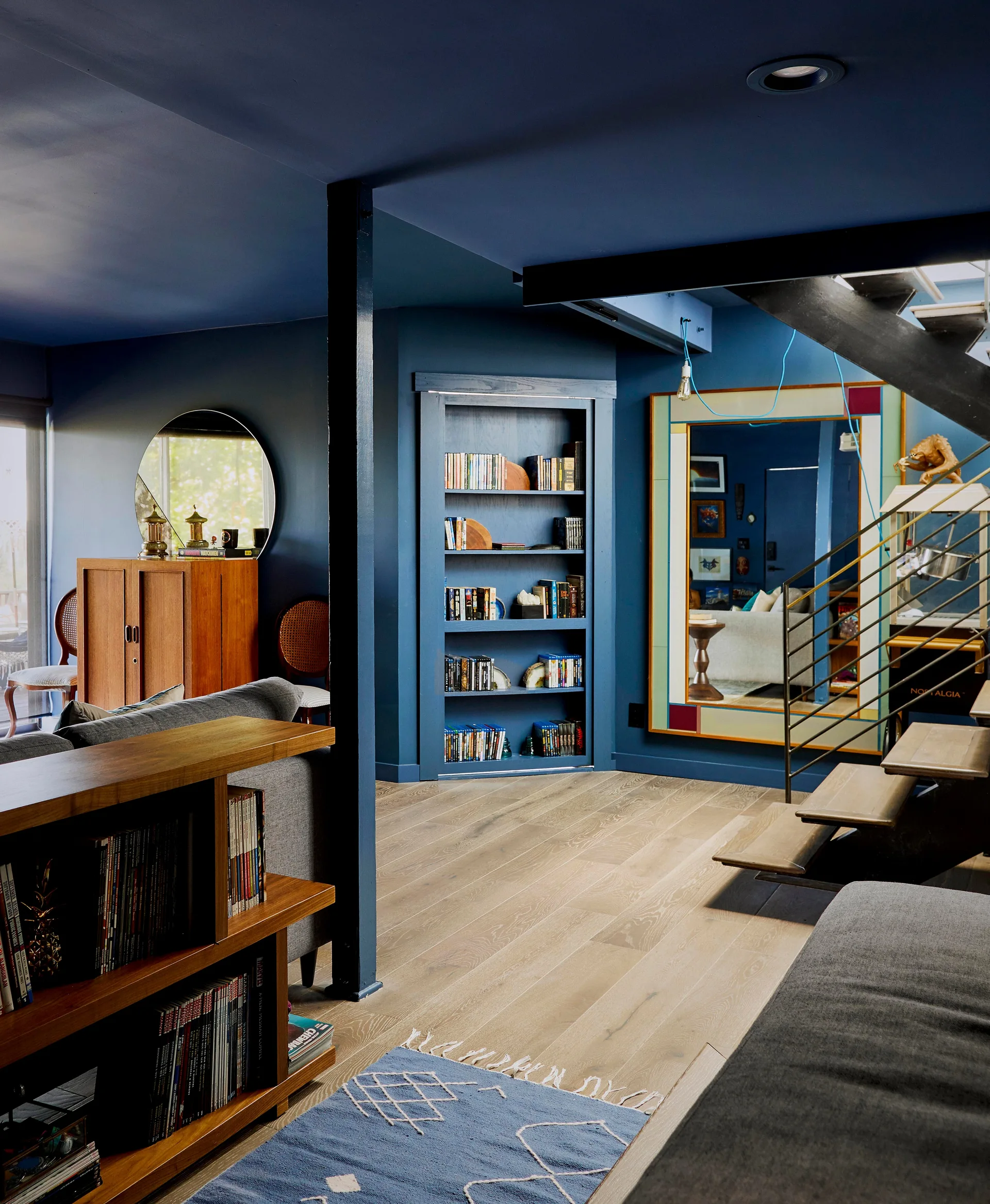 A living room features deep blue walls and ceiling, a built-in bookcase, light wood floors, a grey sofa, and an open-tread staircase.