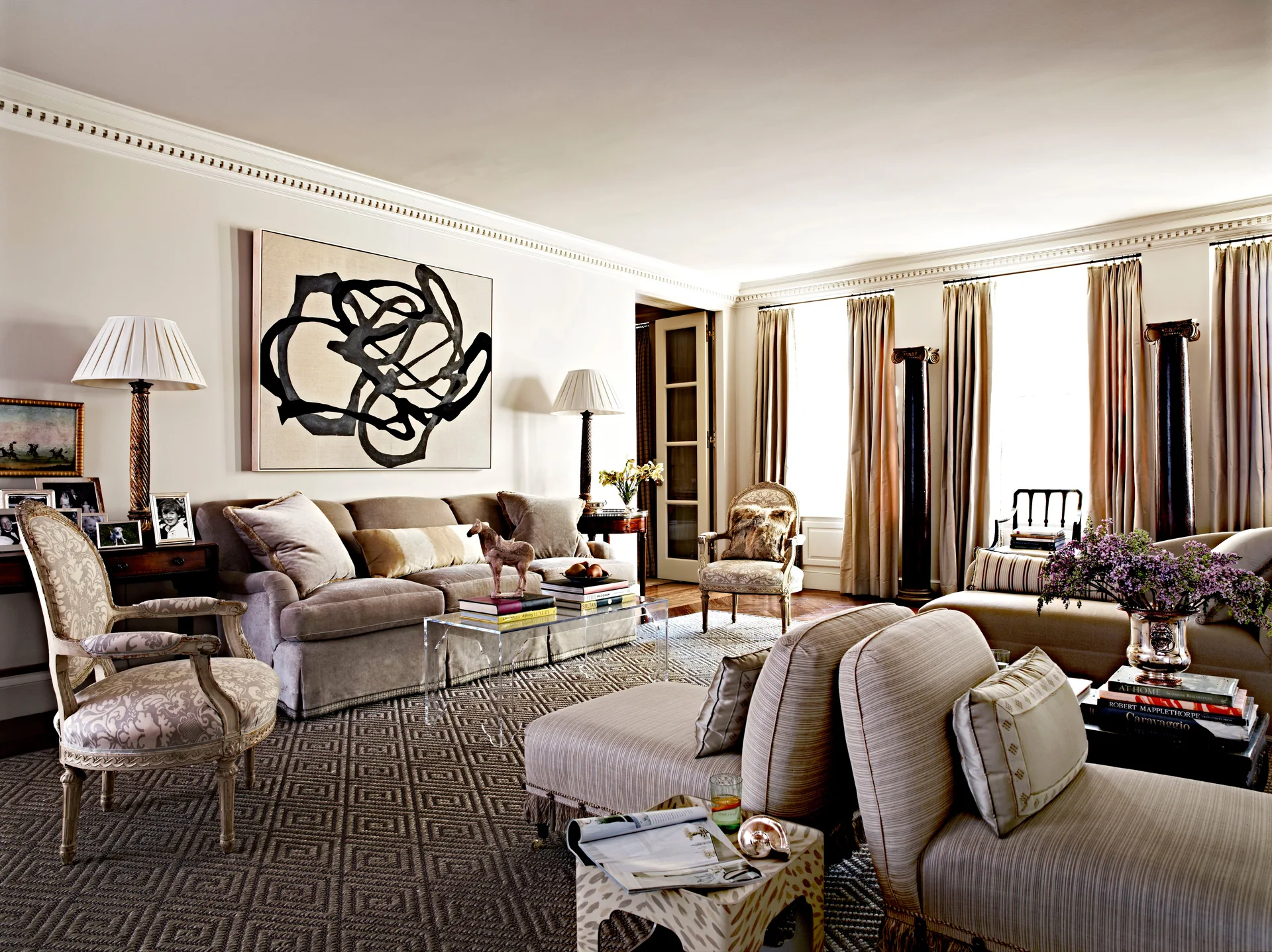 A neutral-toned living room features a beige sofa, large black-and-white abstract painting, patterned geometric rug, and a clear acrylic coffee table.
