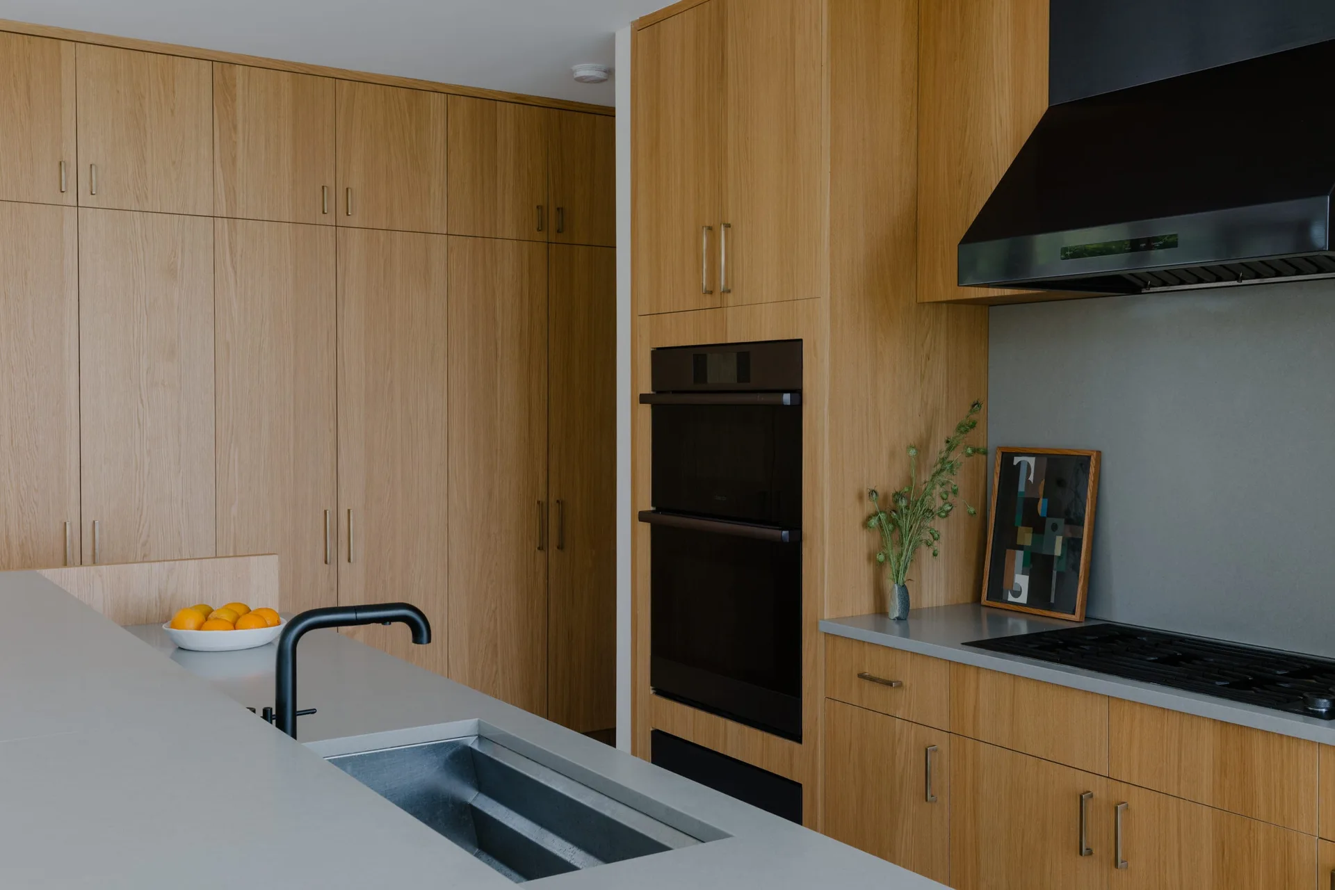A modern kitchen features minimalist flat-panel wood cabinetry, a grey stone island with a black faucet, and an integrated double wall oven.