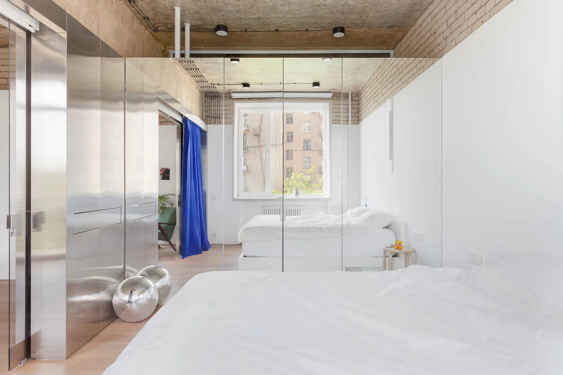 A modern industrial bedroom features a white bed, a large floor-to-ceiling mirrored wall, raw concrete ceiling, and metallic sliding closet doors.
