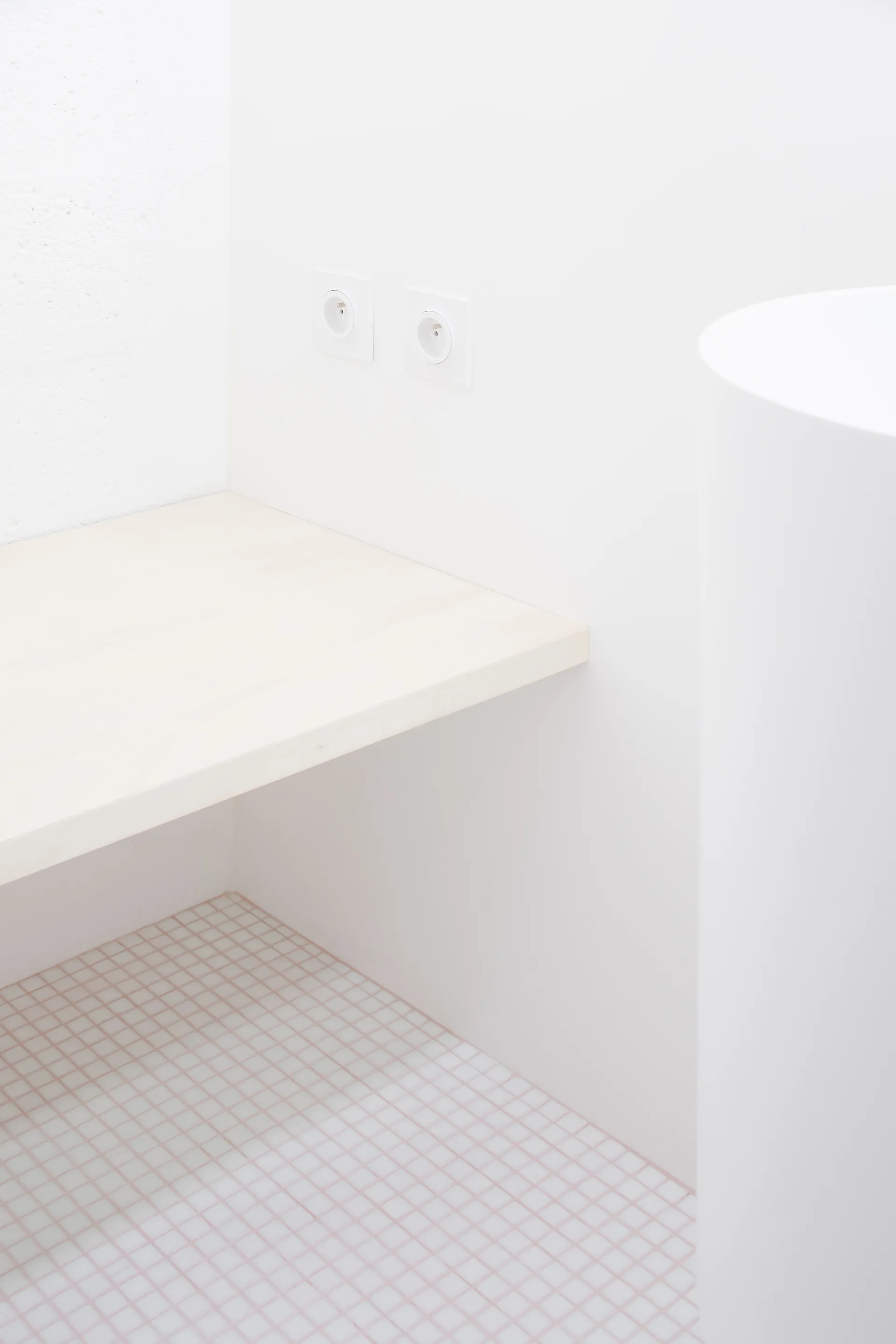 A minimalist white bathroom corner features a light wood floating shelf above a small square grid-tiled floor and two white electrical outlets.