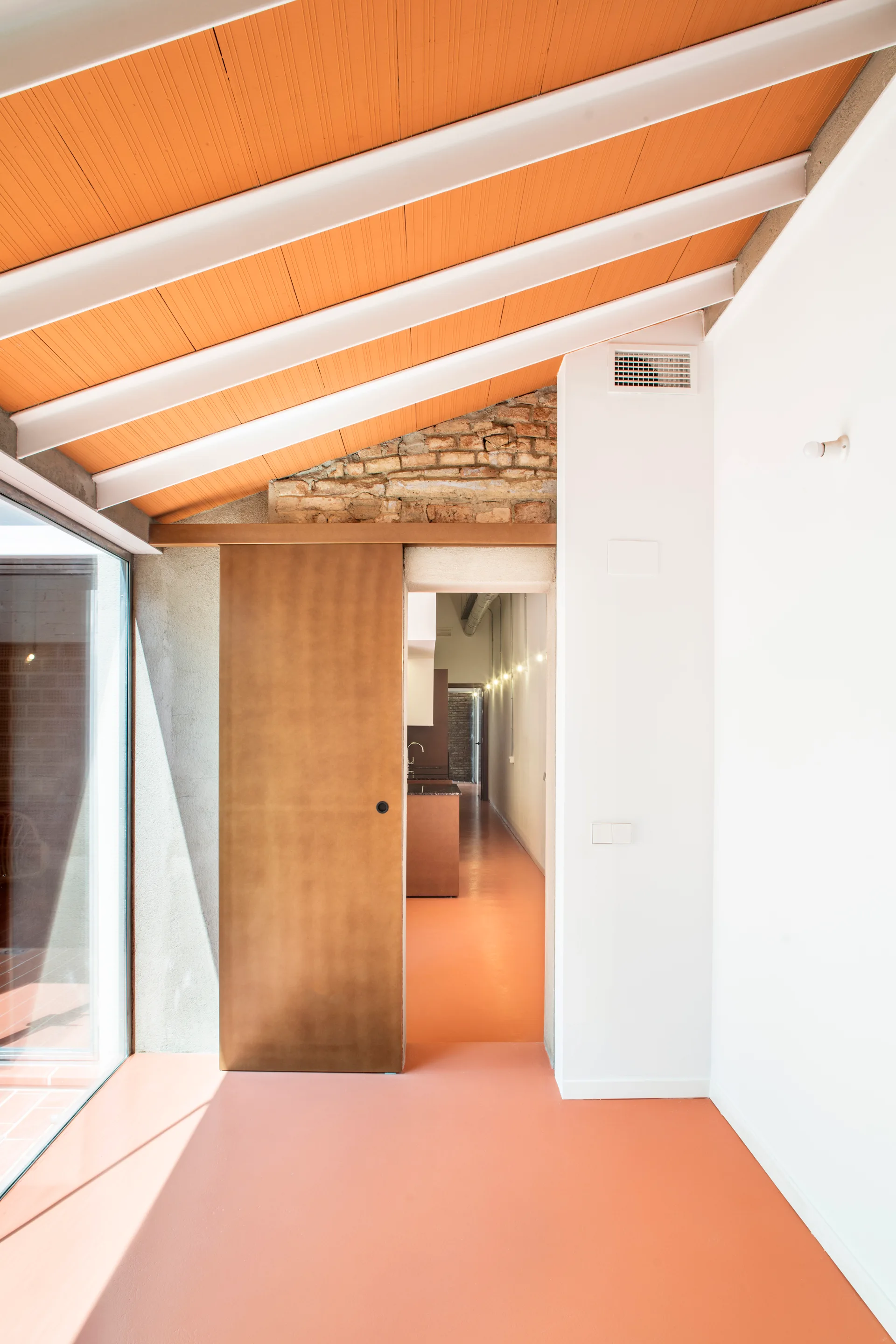 A minimalist hallway features a bright orange floor, white painted ceiling beams, and a sliding wood-toned door leading to an interior corridor.