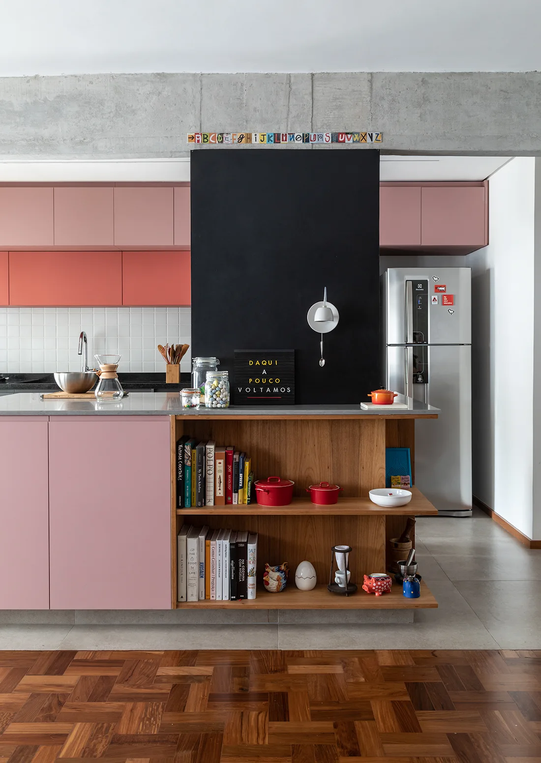 A modern kitchen features pink and coral cabinetry, an exposed concrete ceiling beam, and a wooden island with open shelving.