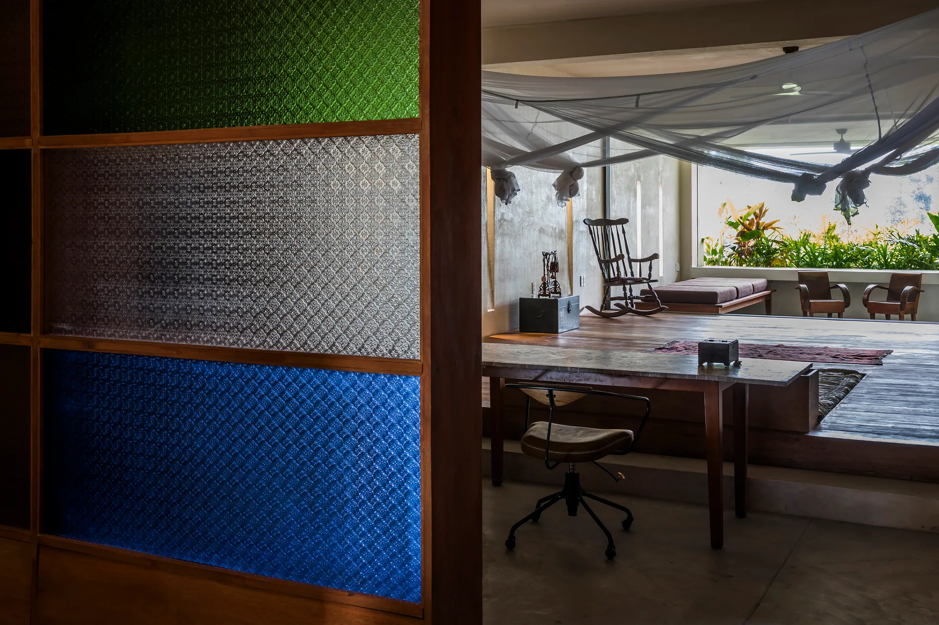 A multi-colored patterned glass screen partitions a workspace desk from a raised wooden platform living area with seating and a white canopy.
