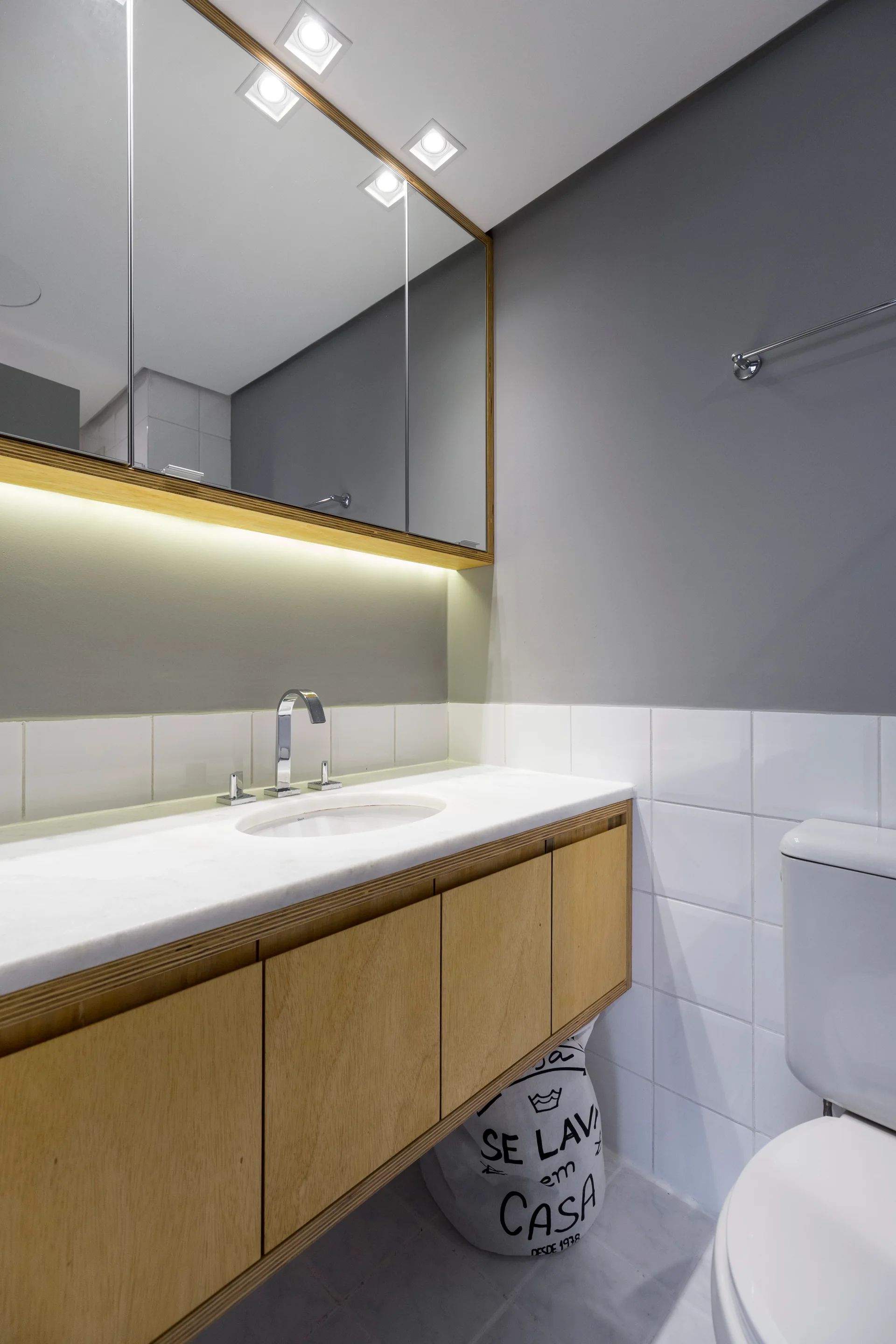 A floating light wood vanity sits below a large mirrored cabinet with integrated lighting in a gray bathroom with white grid tile.