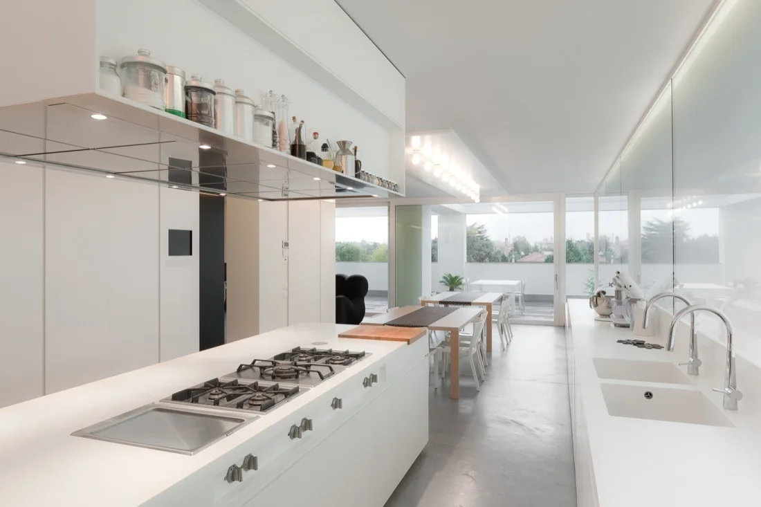 A minimalist white kitchen features a large central island with a gas cooktop, an integrated sink, and an open shelf holding glass storage jars.