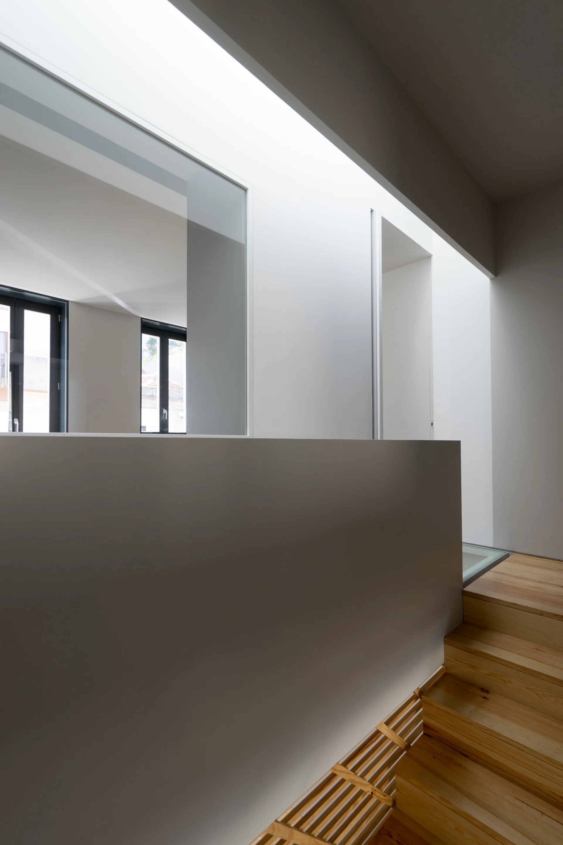 A modern wooden staircase ascends toward a landing lit by a large skylight, featuring smooth grey wall partitions and black-framed windows.