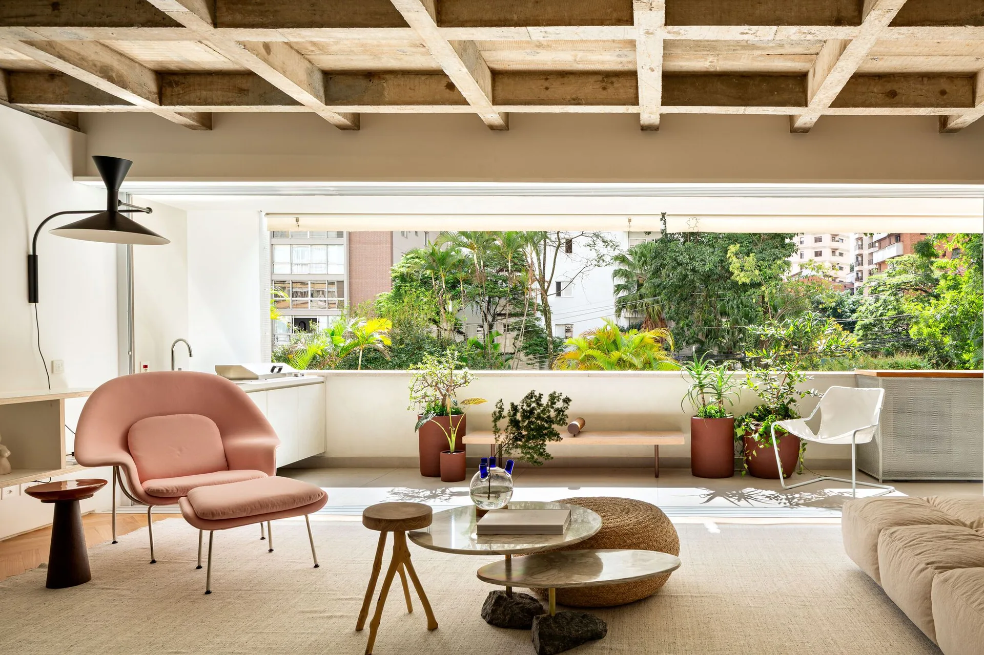 A bright living room features a pink armchair, concrete coffered ceiling, and glass sliding doors opening to a balcony with potted plants.