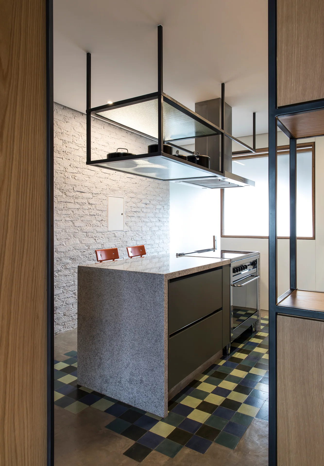A modern kitchen features a stone-wrapped island with a built-in stove, an overhead black metal shelf unit, and a multicolored checkerboard tile floor.