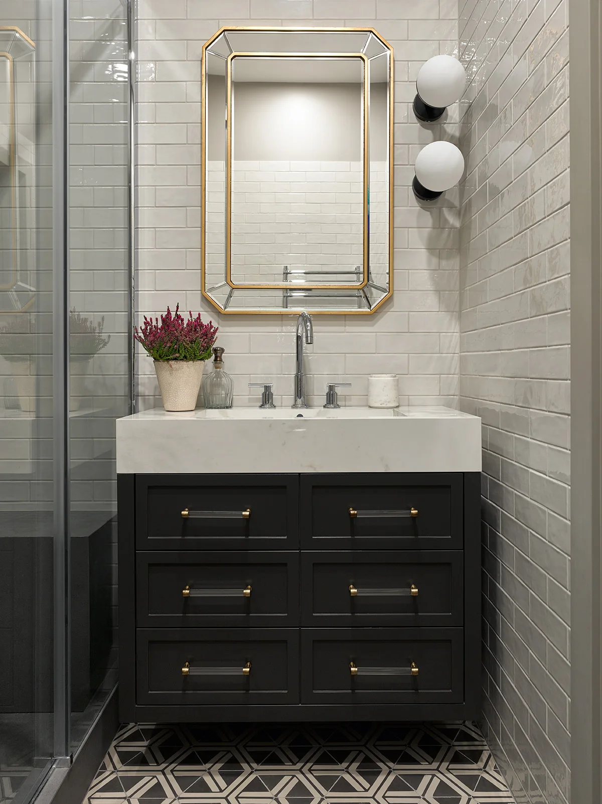 A modern bathroom features a black vanity with a white countertop under a gold-framed mirror, flanked by globe sconces and white subway tiles.