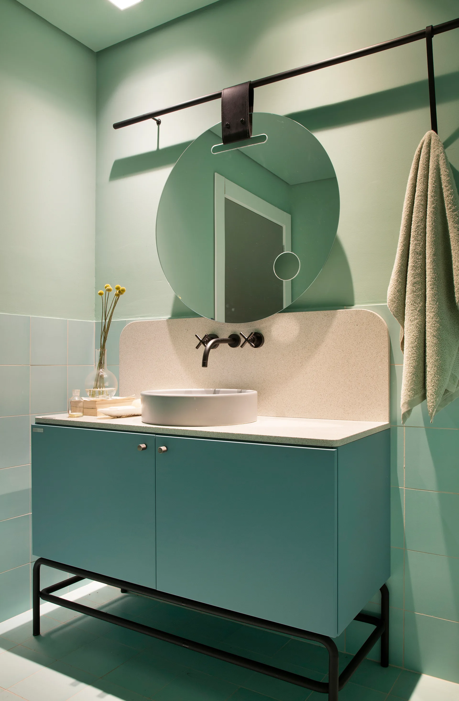 An aqua blue bathroom vanity features a vessel sink, a wall-mounted black faucet, and a round mirror hanging from a black metal rail.