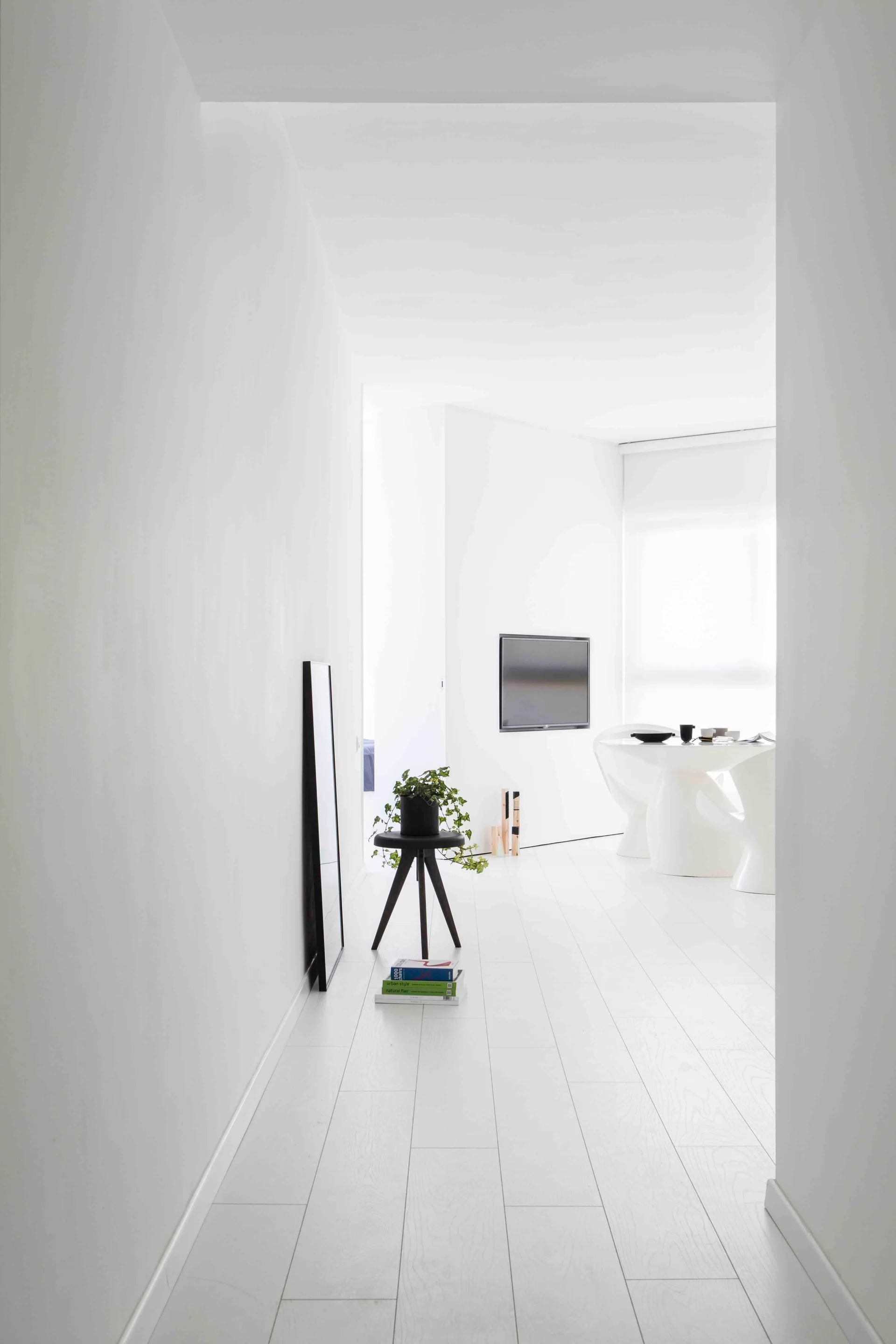 A bright minimalist white hallway features a black tripod table with a plant, leading to a dining area with sculptural white chairs and a table.
