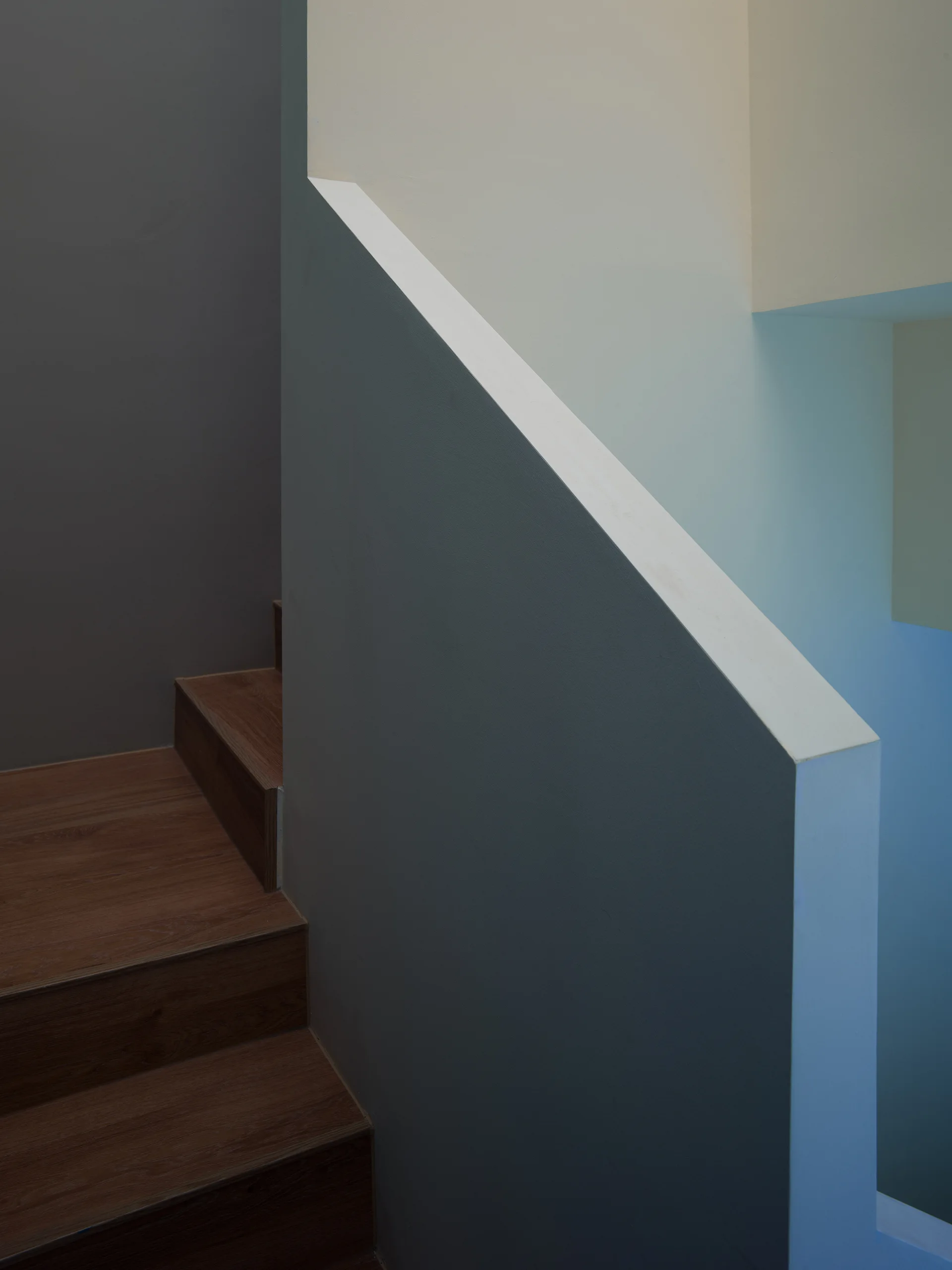 A close-up view of a minimalist wooden staircase featuring dark wood treads and a solid, sharply angled geometric wall painted in gray and white.