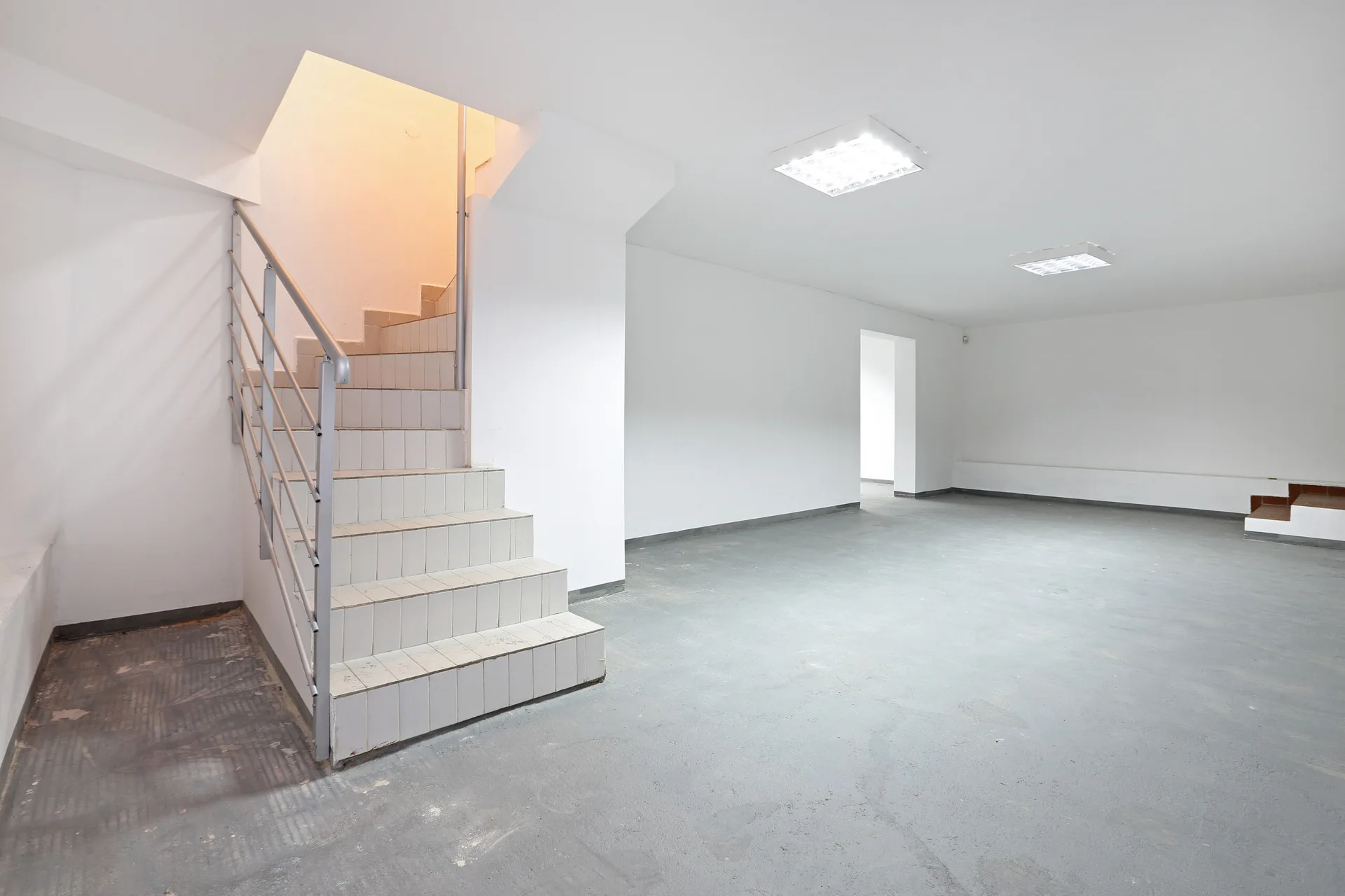 An empty minimalist room featuring white walls, a grey concrete floor, square ceiling lights, and a tiled staircase with a metal railing.