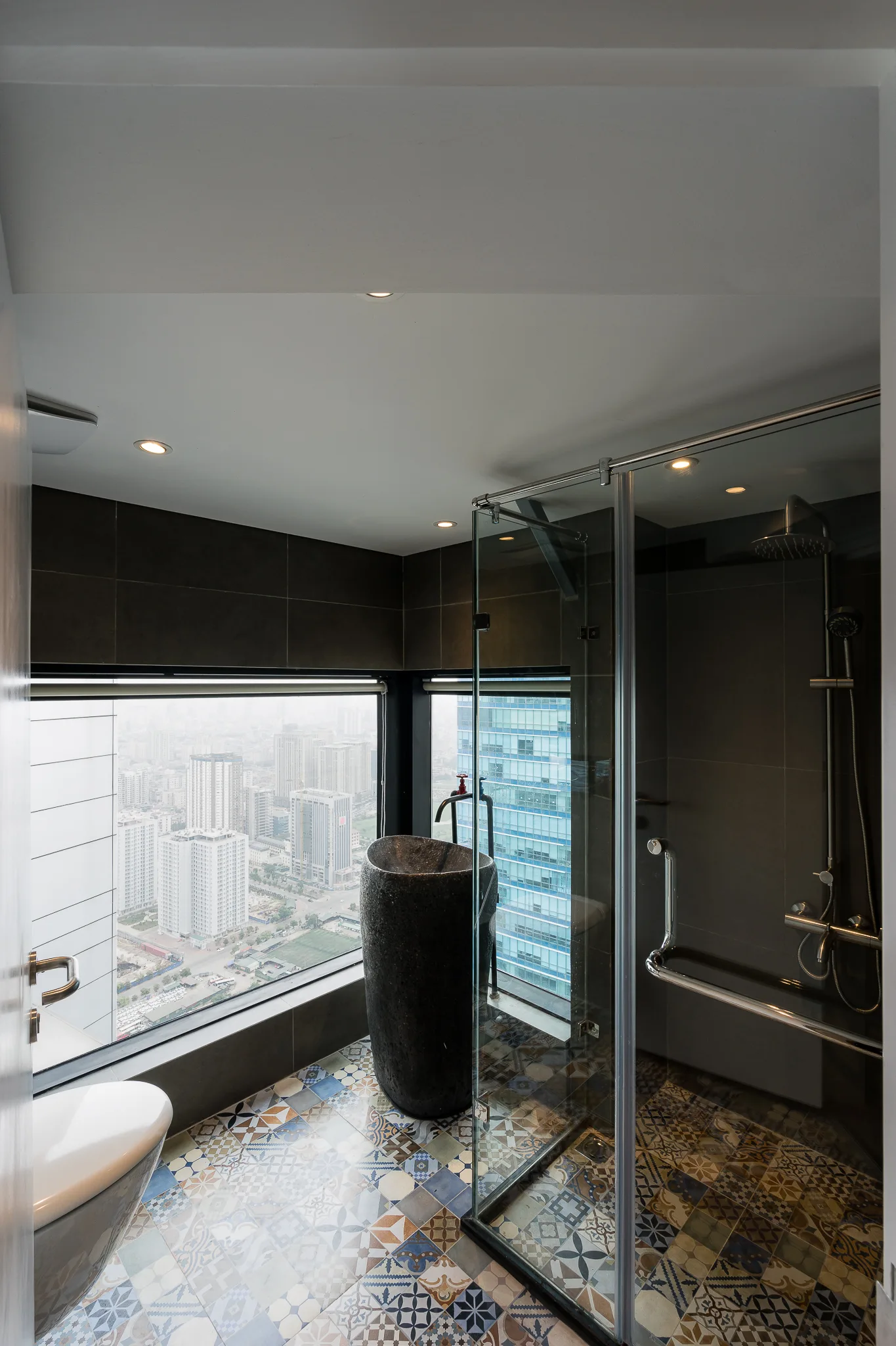 A modern high-rise bathroom featuring a dark stone pedestal sink, a glass shower enclosure, patterned floor tiles, and a large window with a city view.