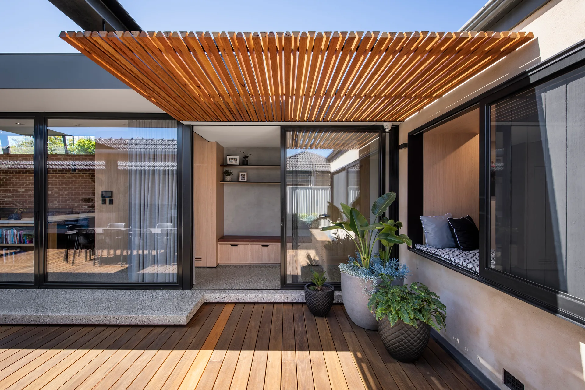 A wooden deck with a slatted timber pergola overhead, featuring large sliding glass doors that open to a modern interior room with built-in cabinetry.